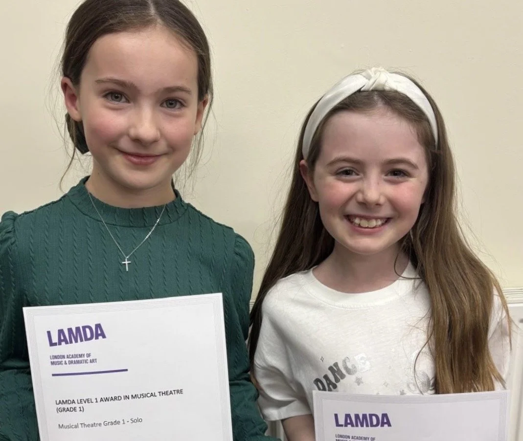 Two young girls smiling and holding awards from the London Academy of Music & Dramatic Art (LAMDA), indicating they received awards in musical theatre for grade 1 solo performance.