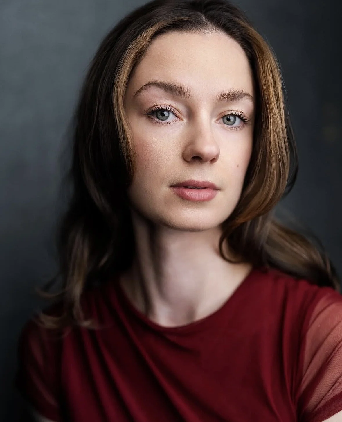 Close-up portrait of a young woman with fair skin, blue eyes, and shoulder-length brown hair styled with soft waves, wearing a red top with sheer sleeves, against a dark background.
