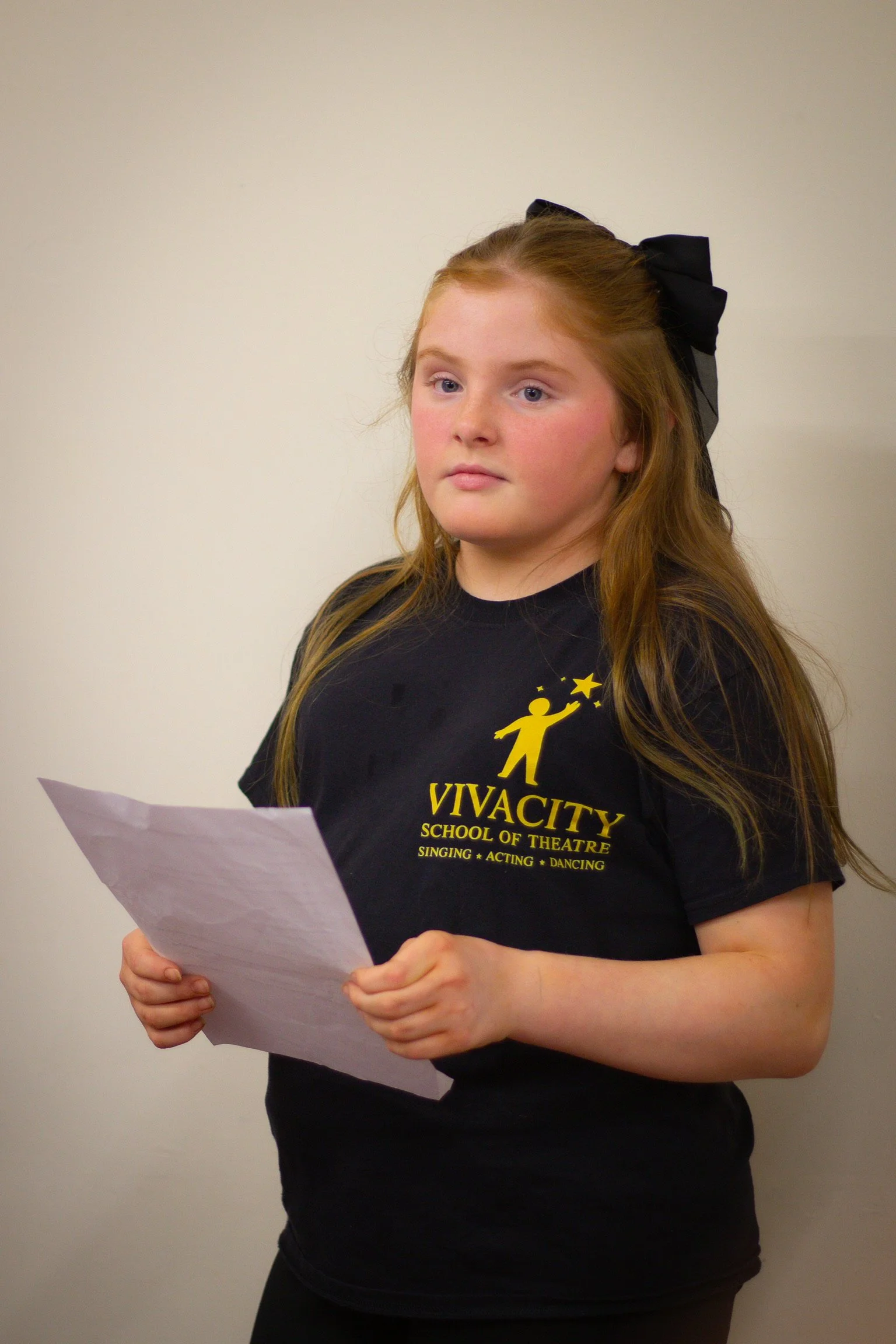 A young girl with red hair, wearing a black t-shirt with yellow text, holding a piece of paper and looking slightly to the side, standing in front of a plain light-colored wall.