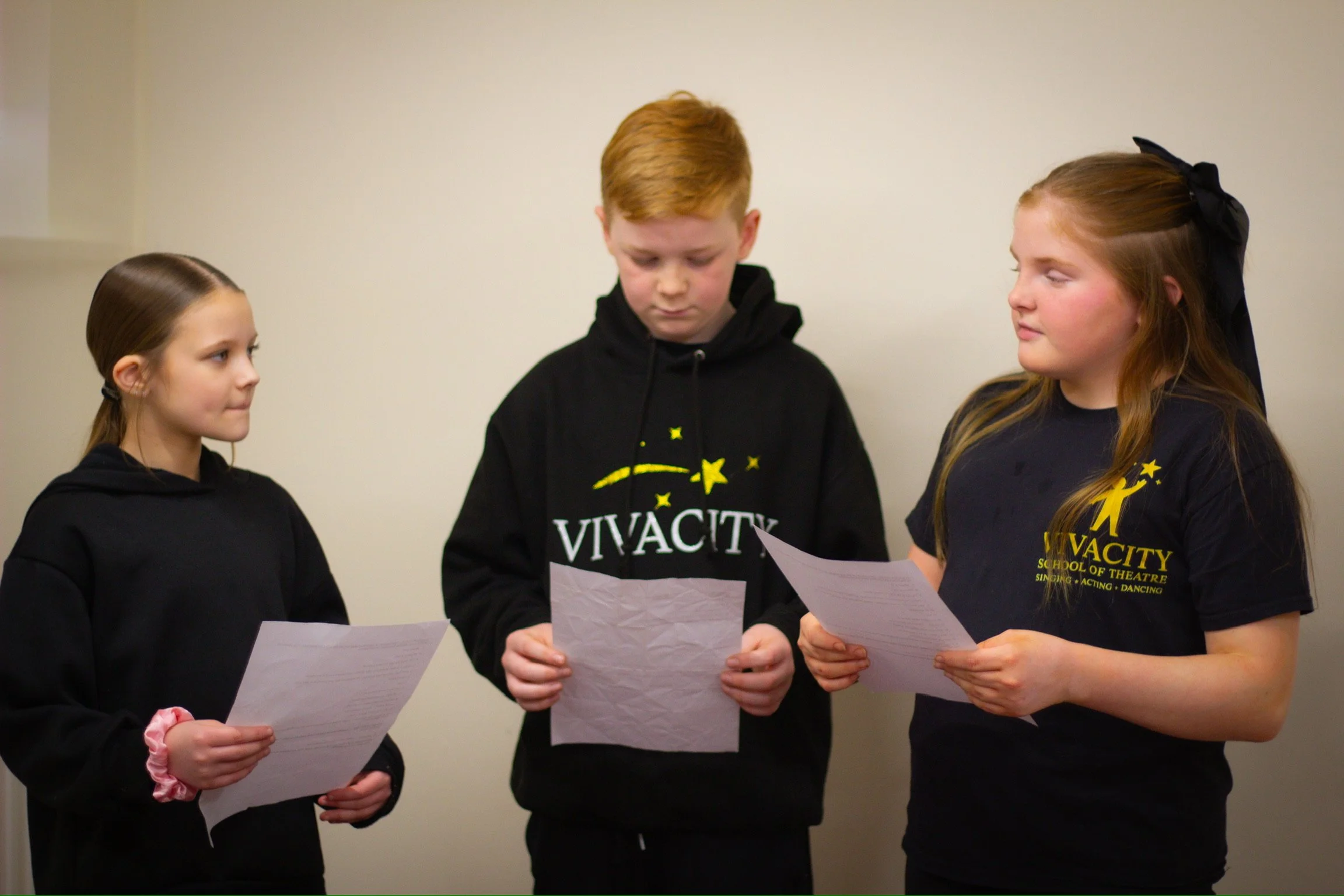 Three children standing together, each holding a piece of paper, possibly reading or singing from it. They are wearing black sweatshirts with yellow and white text related to 'Vivacity School of Theatre', and are in a plain indoor setting with a light-colored wall in the background.
