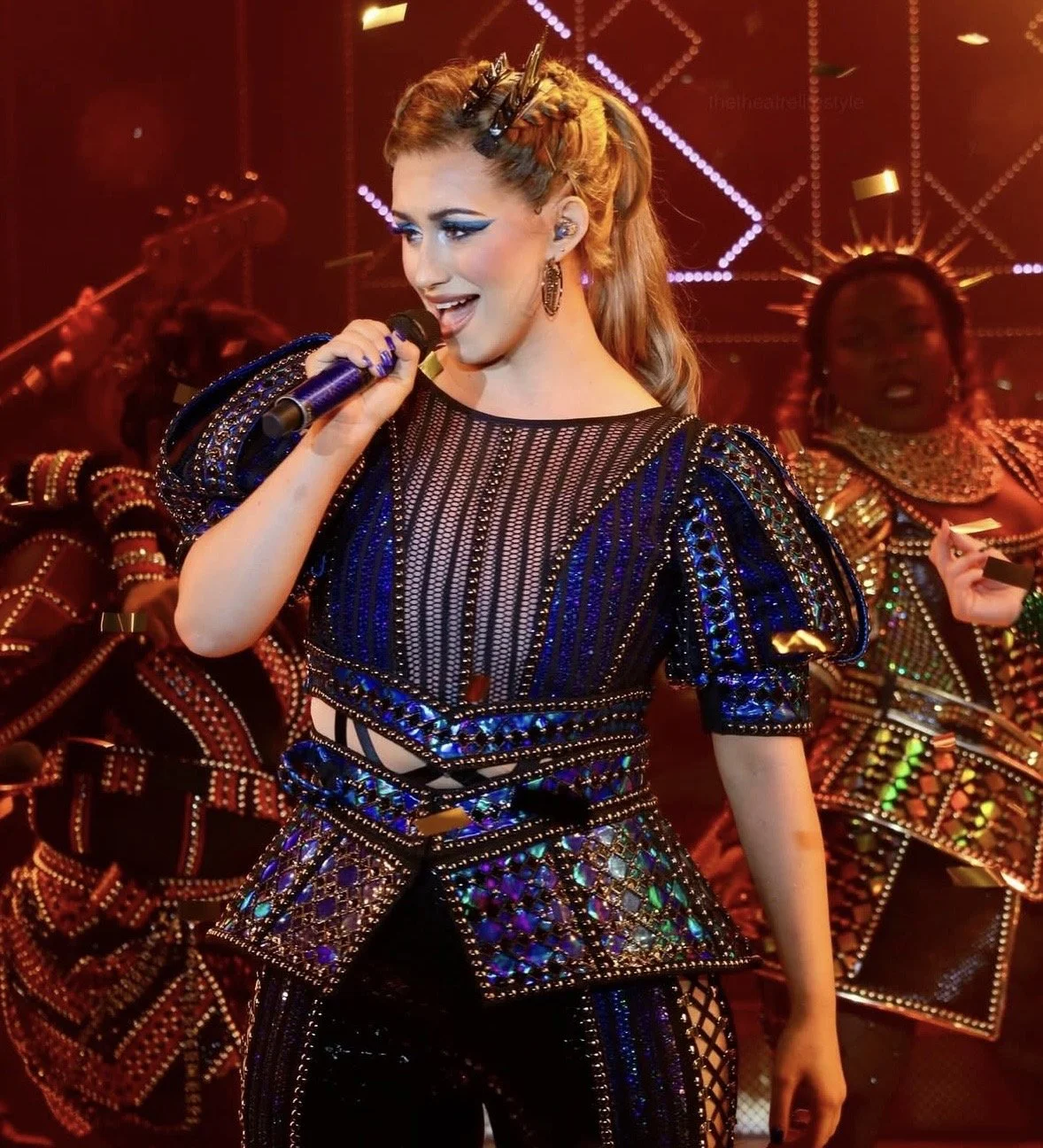 A woman singing into a microphone on stage with vibrant, bejeweled costume and elaborate makeup, surrounded by backup dancers in colorful outfits.