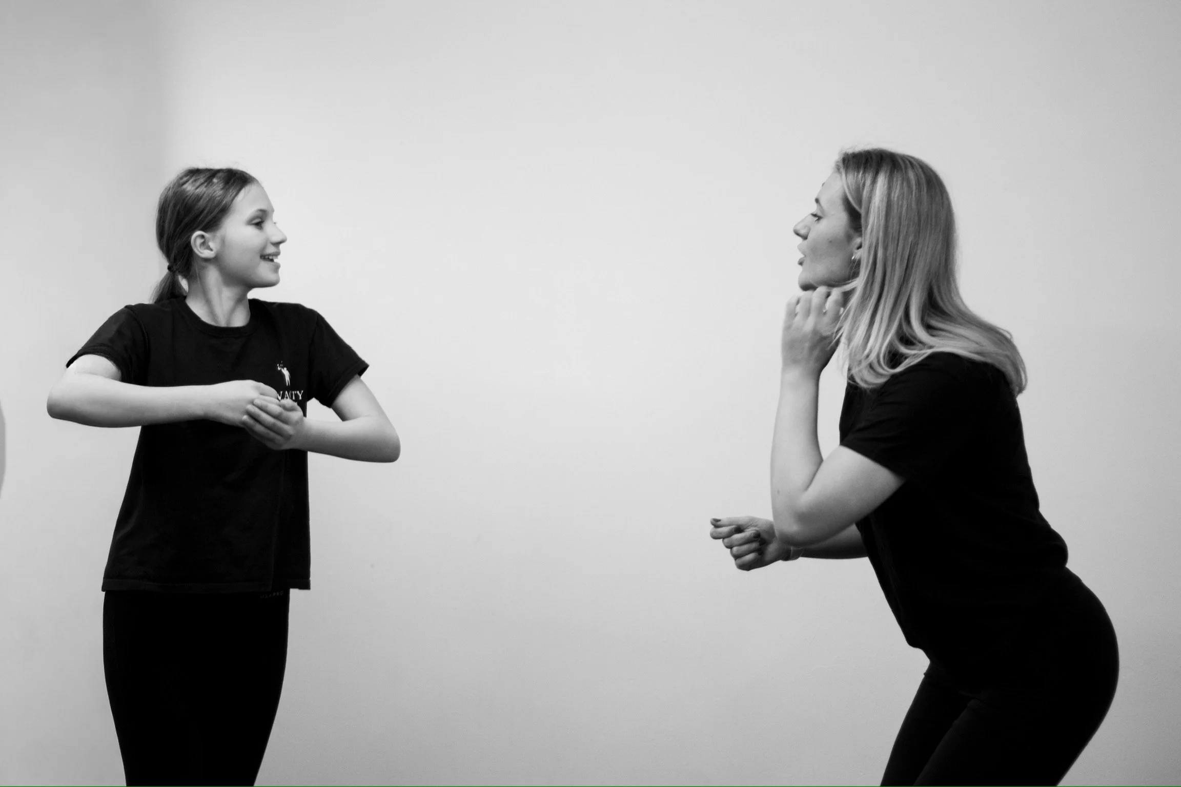 A young girl and an adult woman facing each other, smiling and engaging in a playful or friendly moment, set against a plain background.