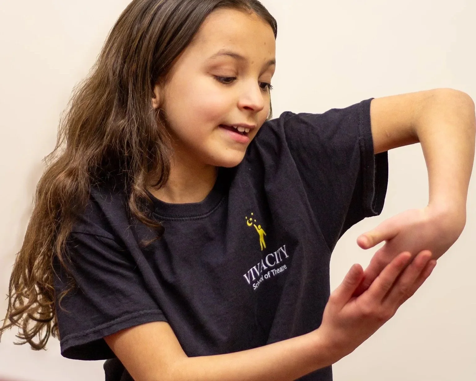 A young girl with long brown hair smiling and looking at her raised left arm, which she is examining. She is wearing a black T-shirt with the logo of Viva City School of Theatre.