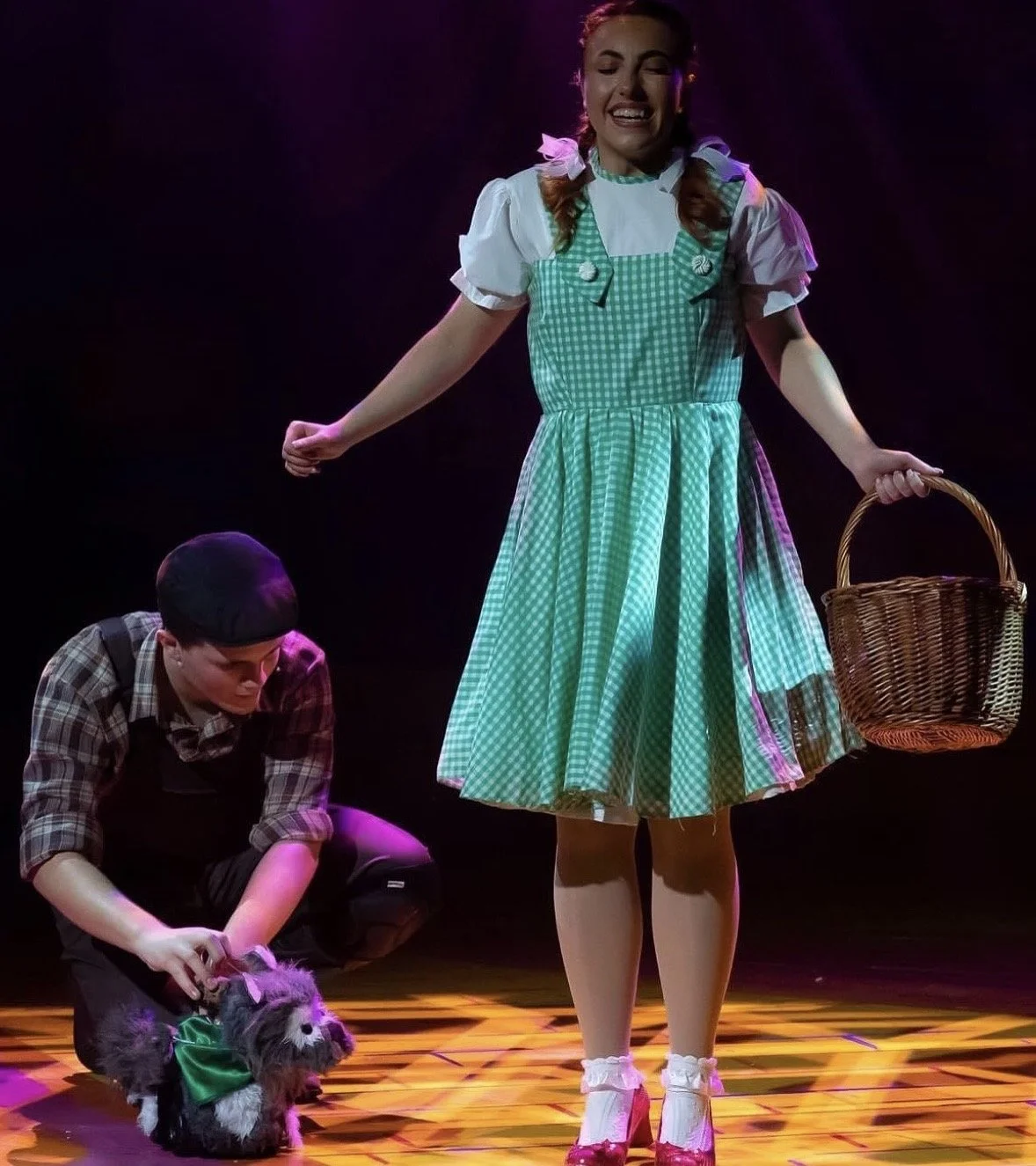 A woman in a green checkered dress with white puffed sleeves standing on stage, holding a wicker basket, with a man kneeling beside her, tending to a small dog with a green bow on its neck. The stage has a colorful patterned floor, and the background is dark with stage lighting.