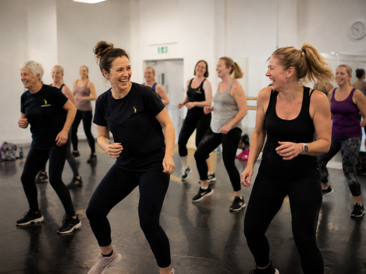 Group of women in athletic wear participating in a dance fitness class in a studio, smiling and enjoying the activity.