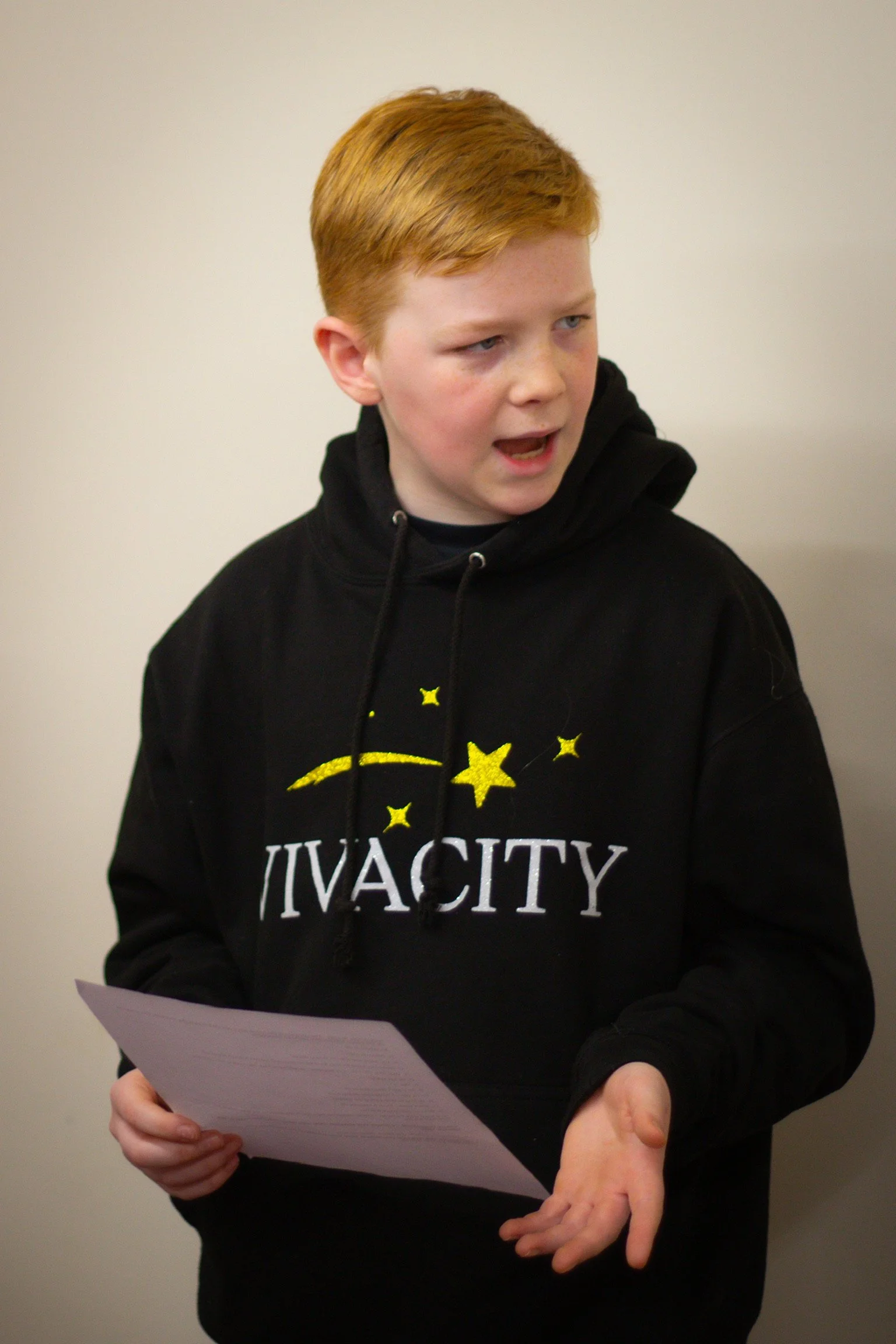 A young person with red hair holding a sheet of paper, wearing a black hoodie with yellow stars and the word 'JIVA CITY' printed on it, standing against a neutral wall.