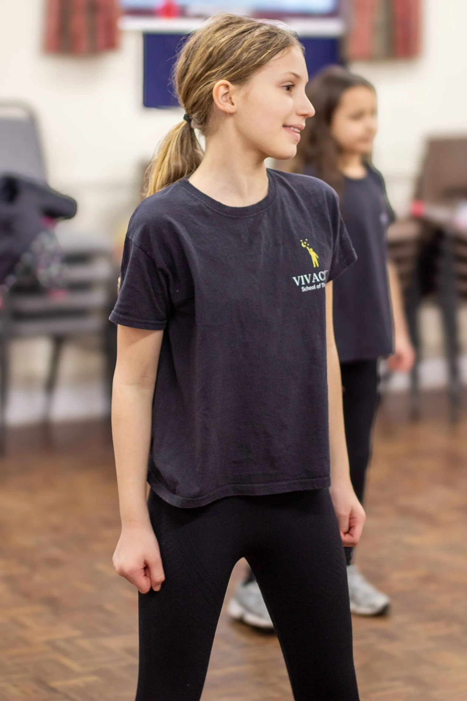 A young girl with light skin, wearing a Vivace School of Theatre t-shirt and black leggings, stands in a dance studio with her right fist clenched, her head slightly turned to the right, and a subtle smile. Other children are visible in the background.