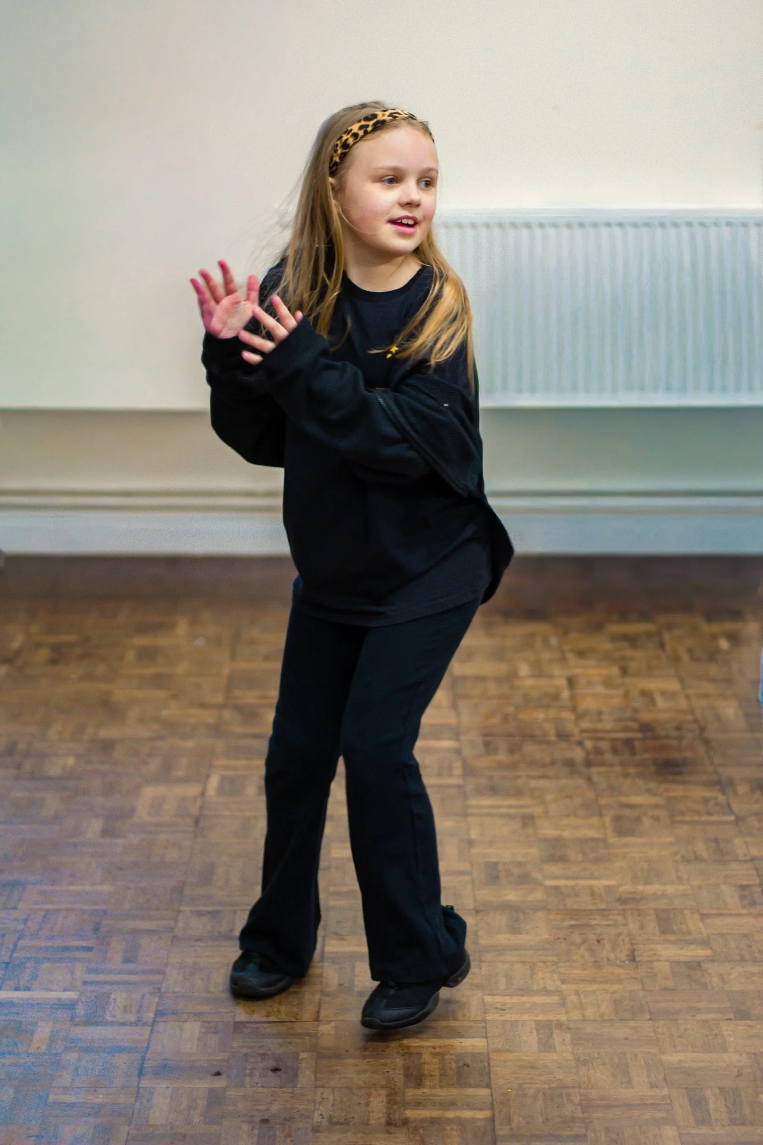 A young girl with long blonde hair, wearing a black outfit and a leopard print headband, standing on a wooden floor with a light-colored wall and radiator in the background, smiling and gesturing with her hands.