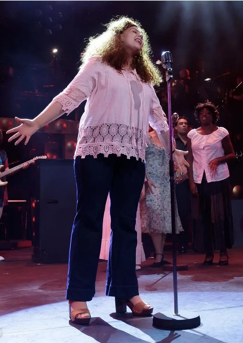 A woman singing on stage with arms outstretched, leading a group of backup singers and musicians in a lively performance.
