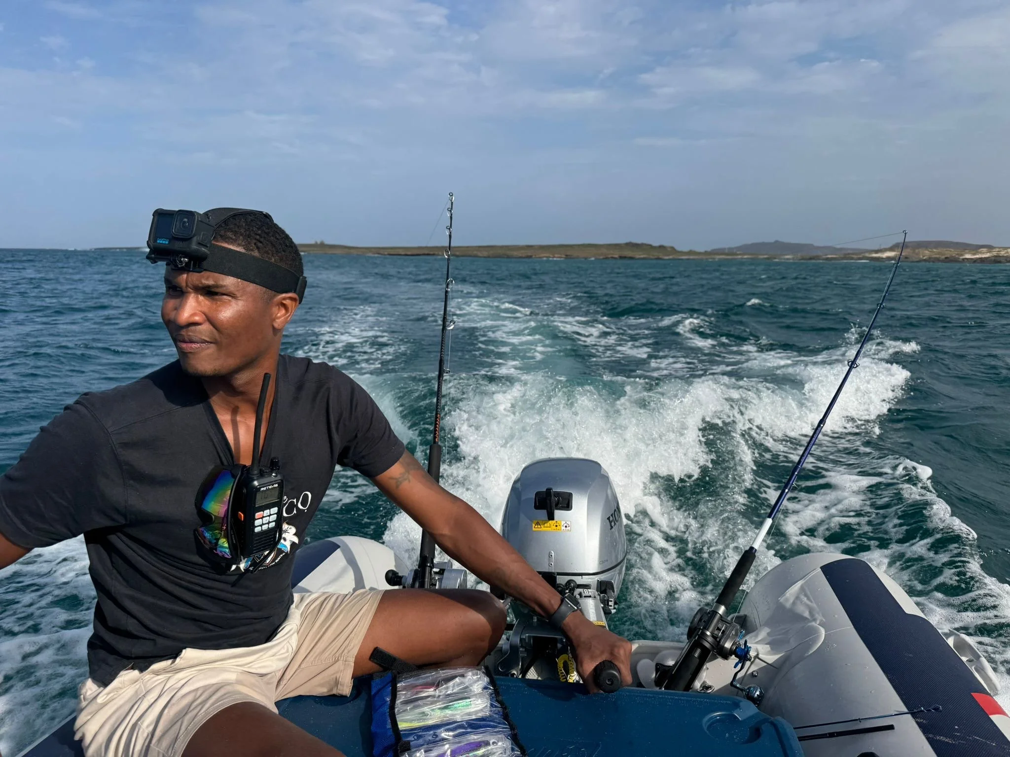 A man is sitting in a boat with two fishing rods, out on the ocean, wearing a black cap with a GoPro camera, a black shirt, and beige shorts. The boat is moving through the water, creating waves behind it, with a distant landmass visible under a partly cloudy sky.