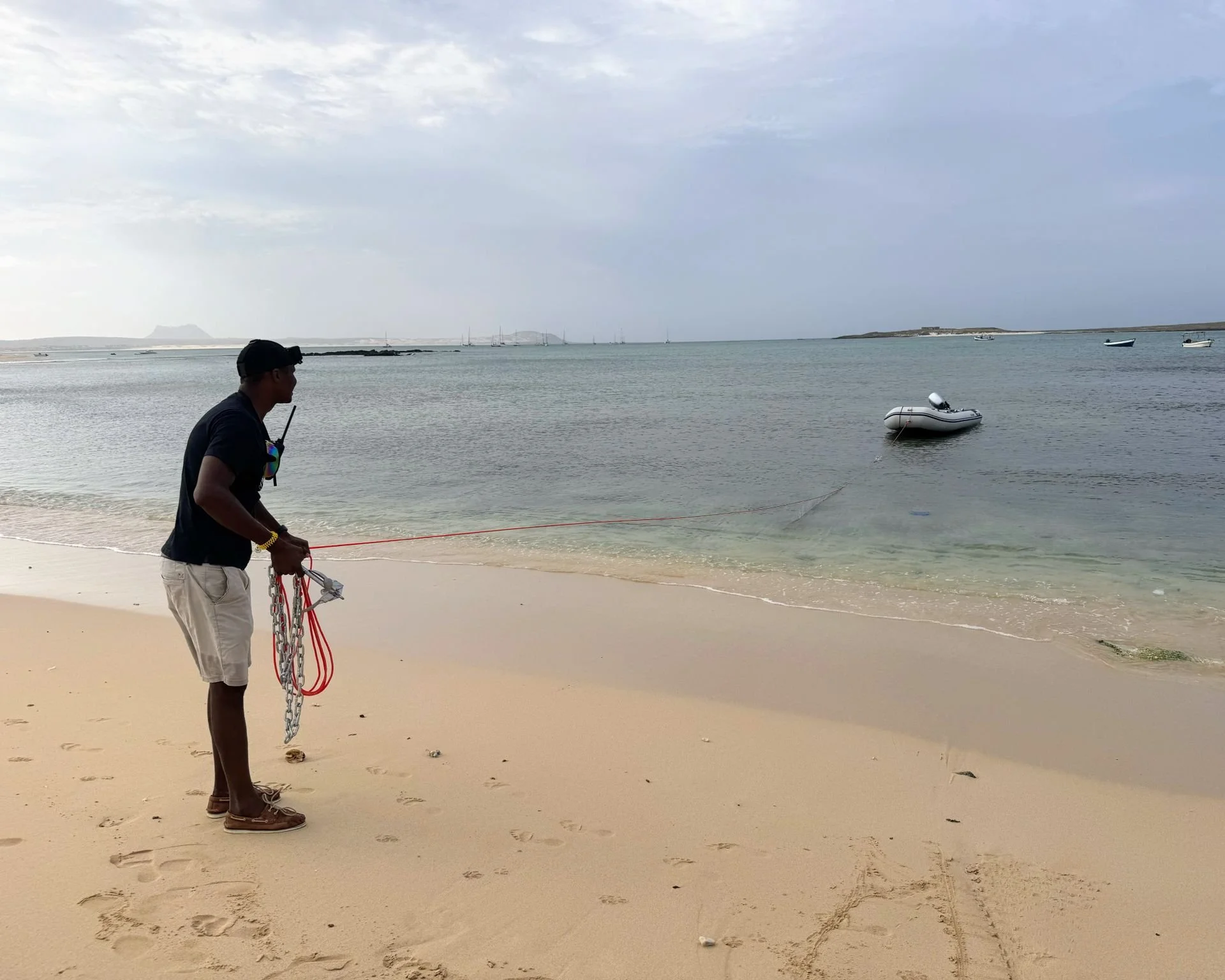 A man holding a chain and a flotation device on the sandy beach, with the ocean, anchored boats, and a cloudy sky in the background.