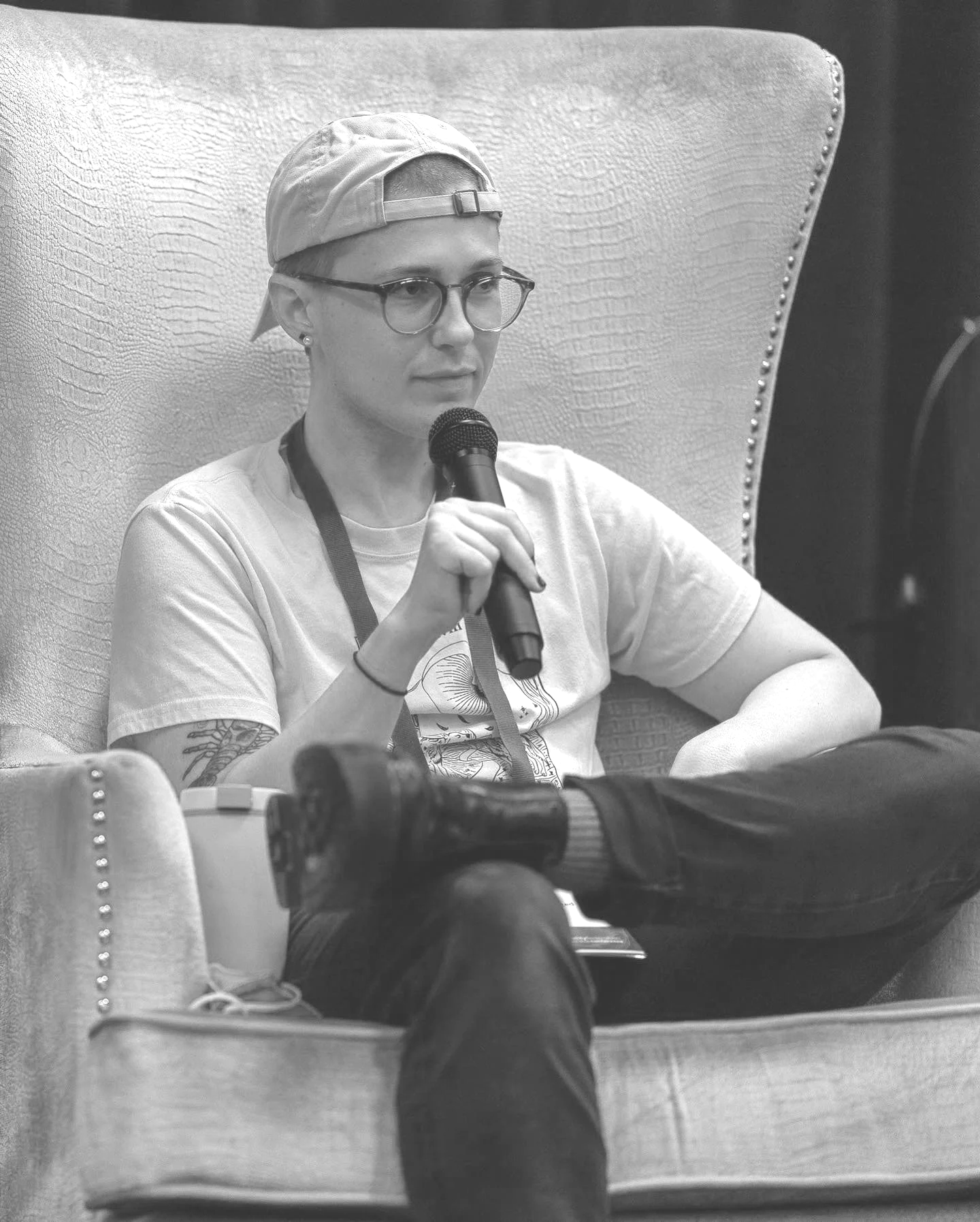 A young white man wearing glasses, a backwards baseball cap, and earrings, sitting in a large chair, holding a microphone, with a tattoo visible on his arm, in a black and white photo.