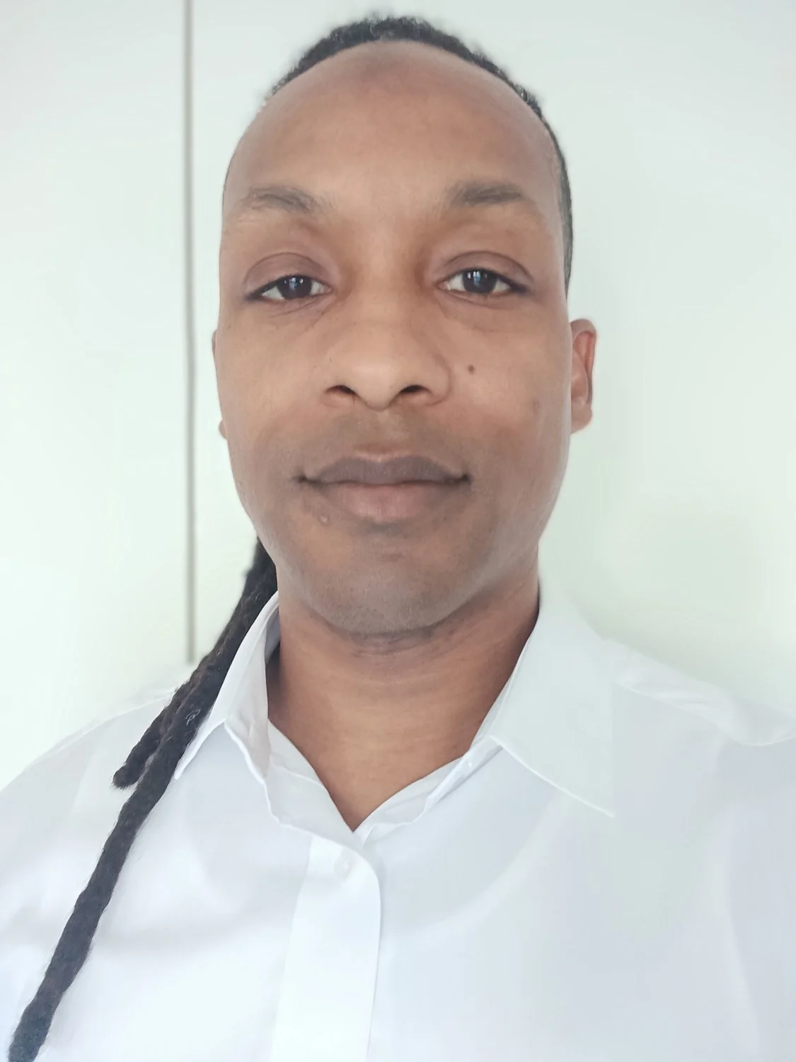 Close-up of a man with dark skin, black braided hair, and a white collared shirt, standing indoors against a light background.