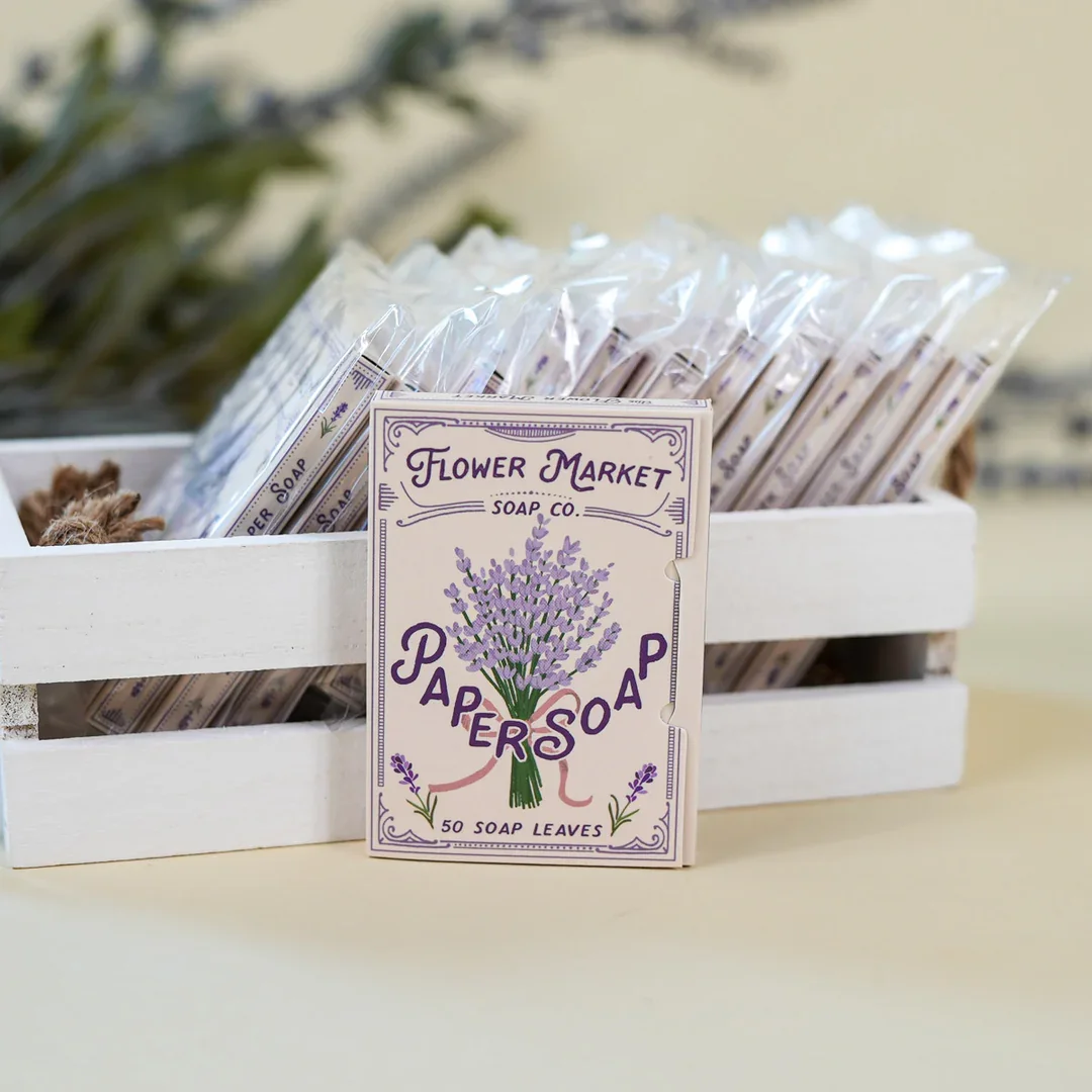 Een wit houten doos met verpakte zeepblokjes en een label dat 'Flower Market Paper Soap' en een bloemenafbeelding van lavendel toont.