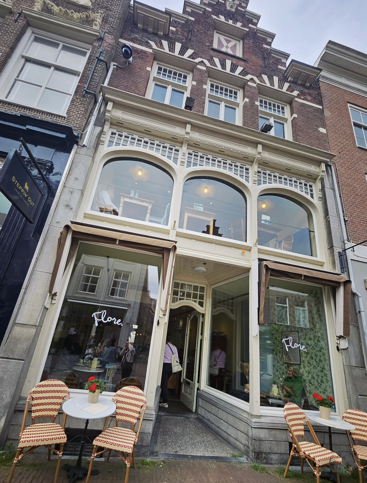Gevel van een café genaamd 'Flore' met een terras met drie ronde tafels en vier stoelen, ramen met houten luifels, en mensen binnen en buiten.