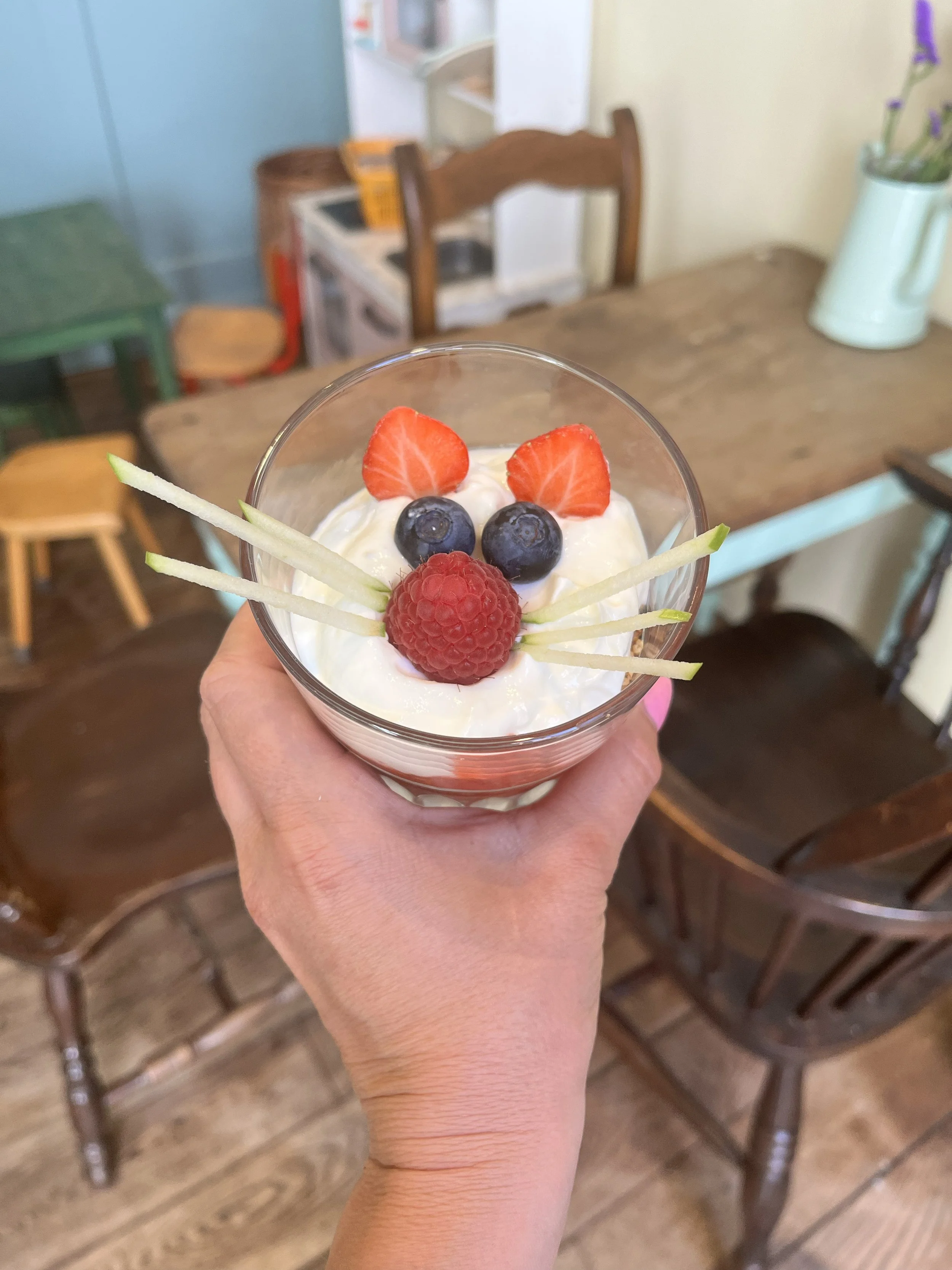 Een hand houdt een glas met een dessert dat versierd is met aardbeien, blauwe bessen, frambozen en dunne stukjes appel. Op de achtergrond zijn houten stoelen en een tafel zichtbaar.