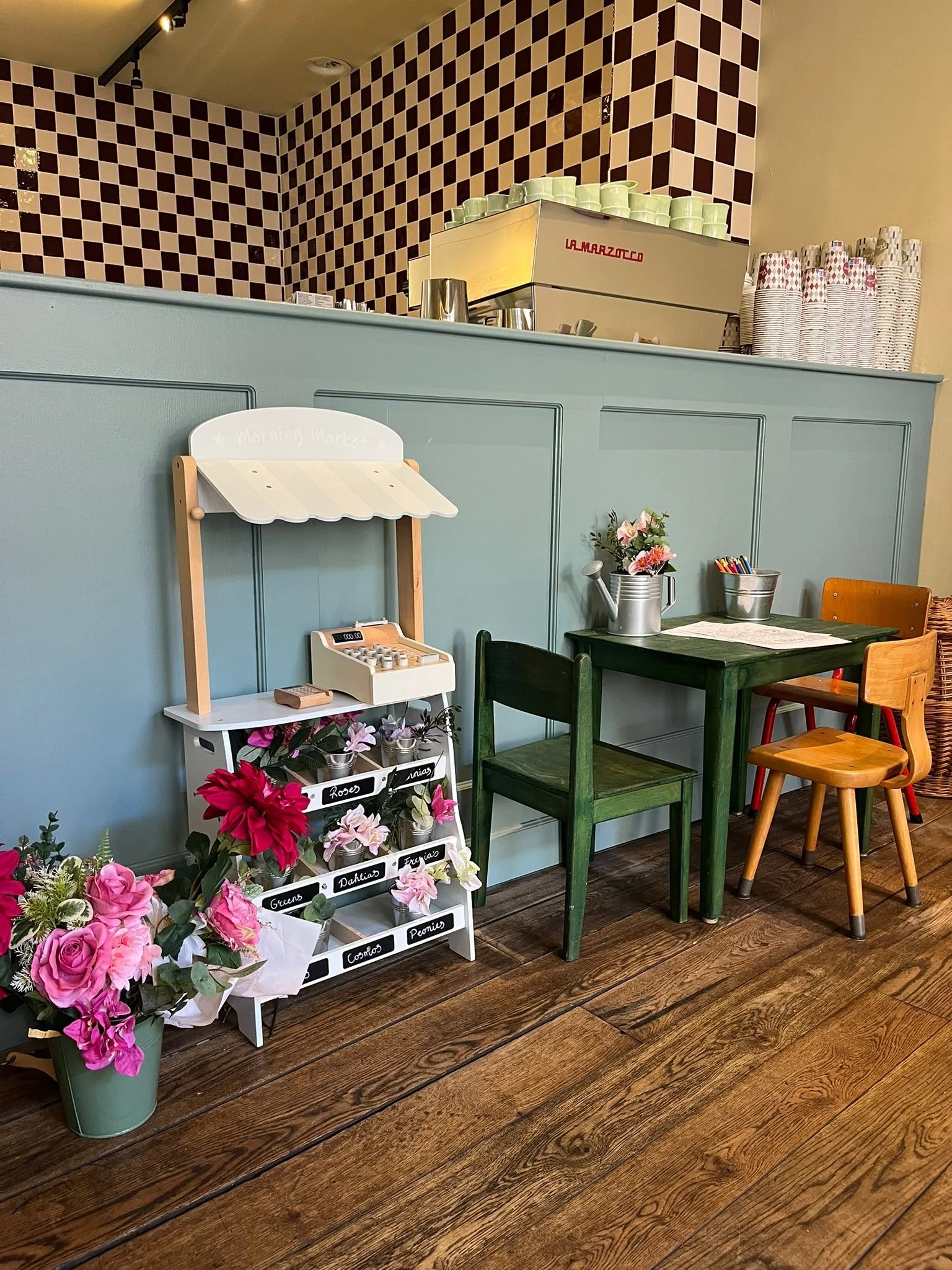 Een interieur met een blauwe houten bar, een bloemenhoek met roze en paarse bloemen, en een muur met een patroon van rood en beige tegels. Er staan verschillende stoelen rondom een kleine tafel, en er is een wit rek met labels van verschillende bloemen.