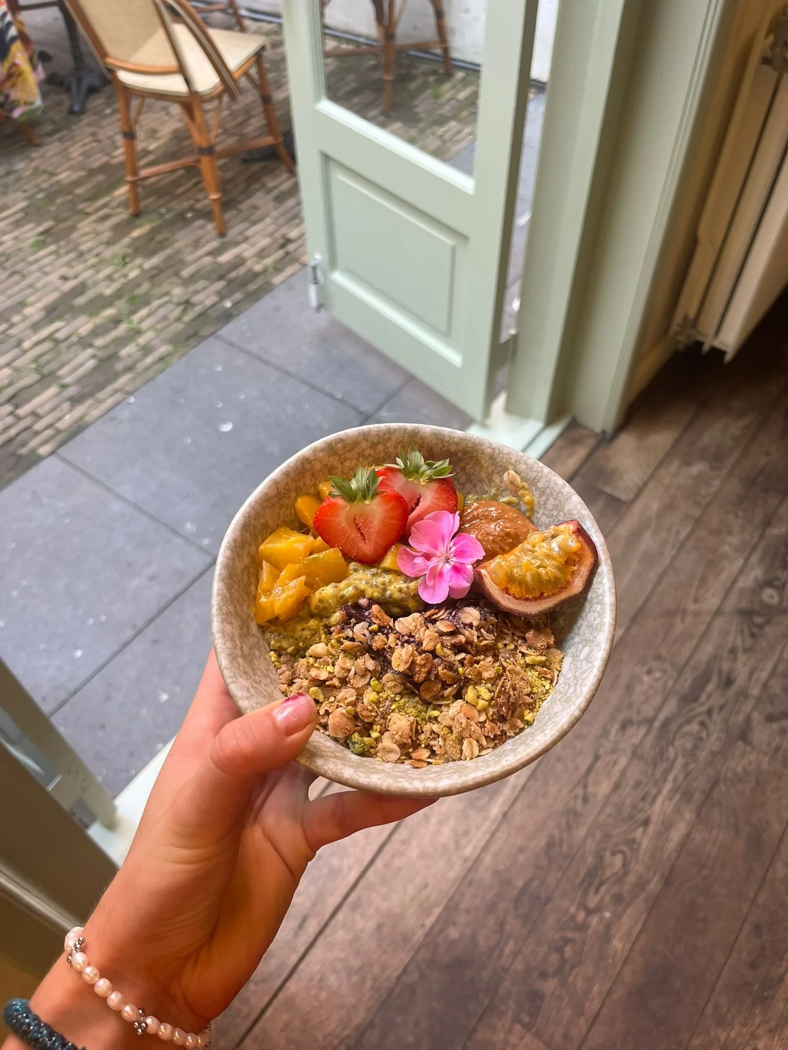 Hand houdt kom met smoothie bowl met aardbeien, mango, passievrucht, granola, en een roze bloemenversiering, binnen voor een open deur.