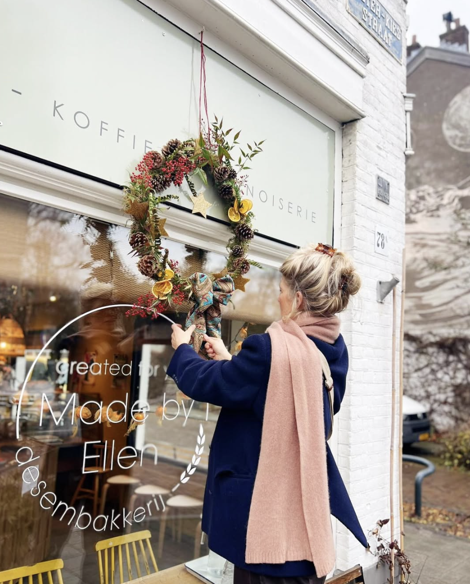Vrouw die een kerstkrans ophangt voor een winkel met een bord dat zegt 'Made by Ellen'.