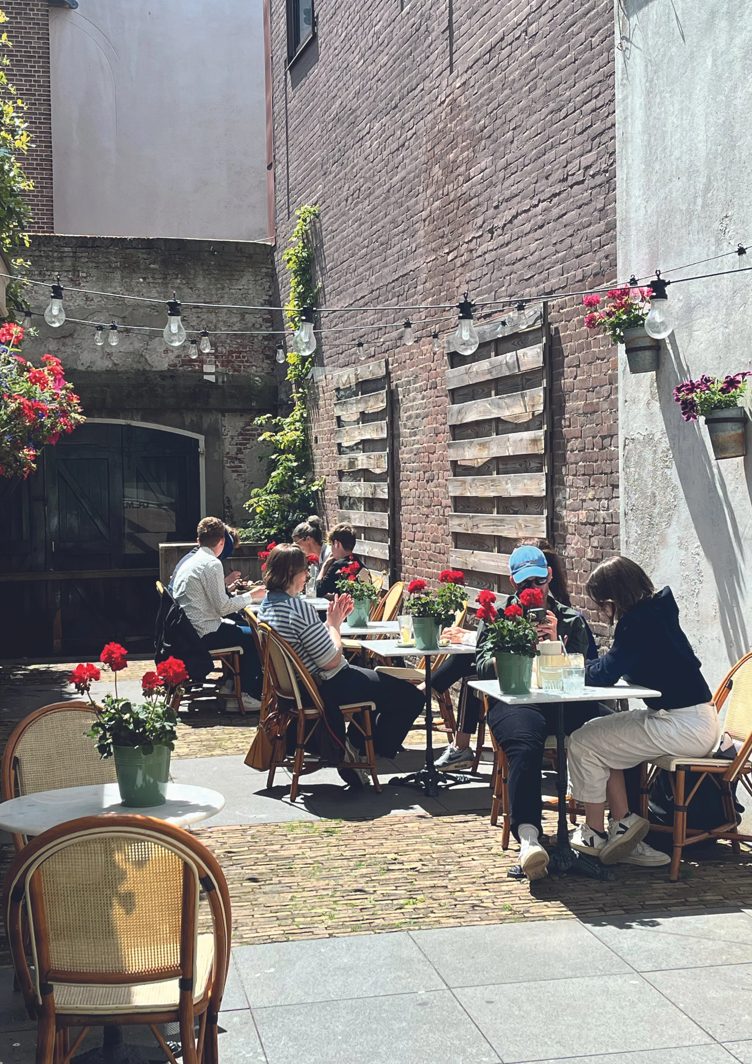 Mensen zitten buiten op terrastafels met bloemen, omgeven door muren met hanglampen en planten.