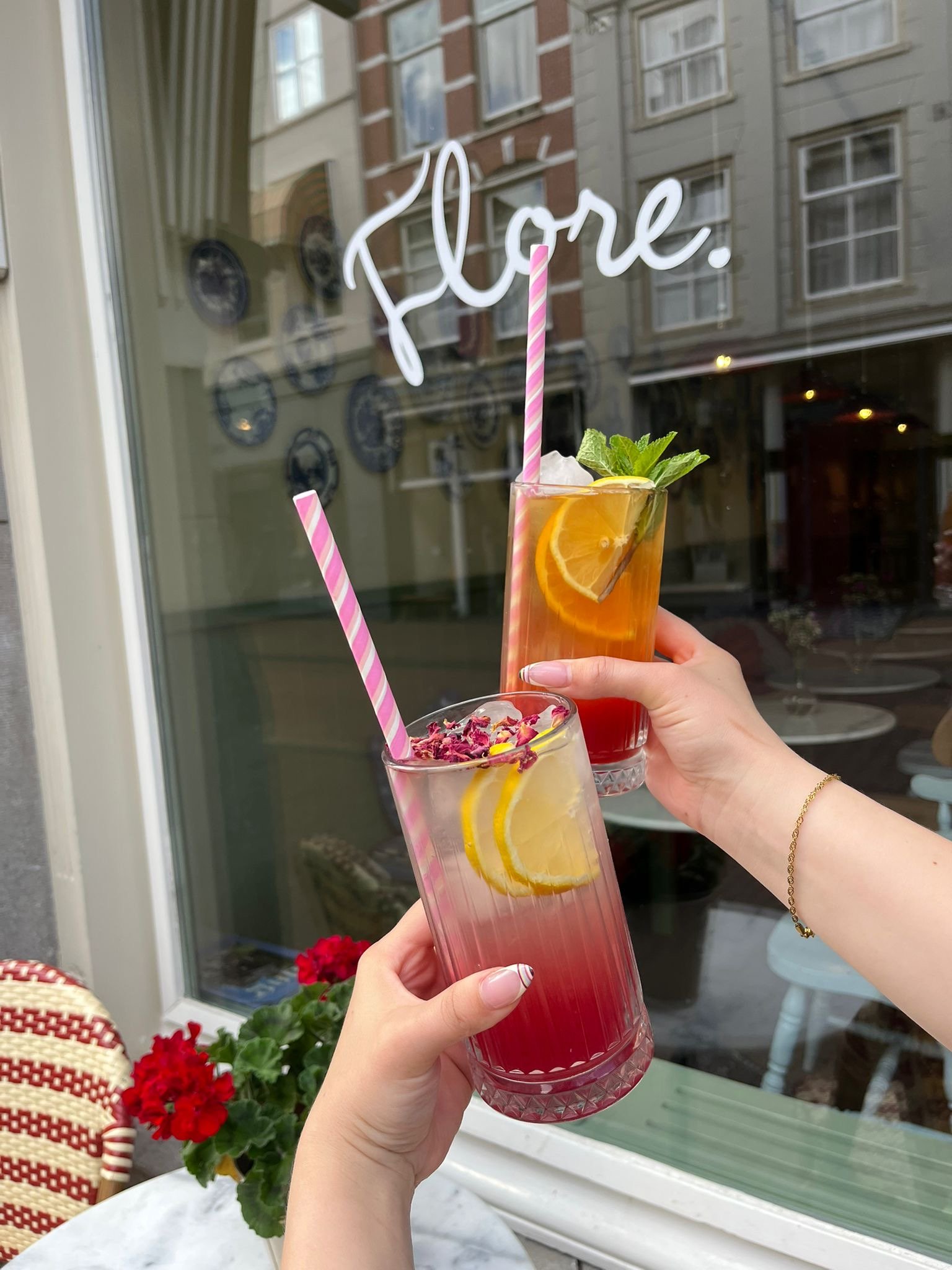Een hand houdt een glas met een rode drank, citroen, munt en ijs, met een roze-wit gestreepte rietje. Op de achtergrond staat een raam met een beschrijving: 'Enjoy'.