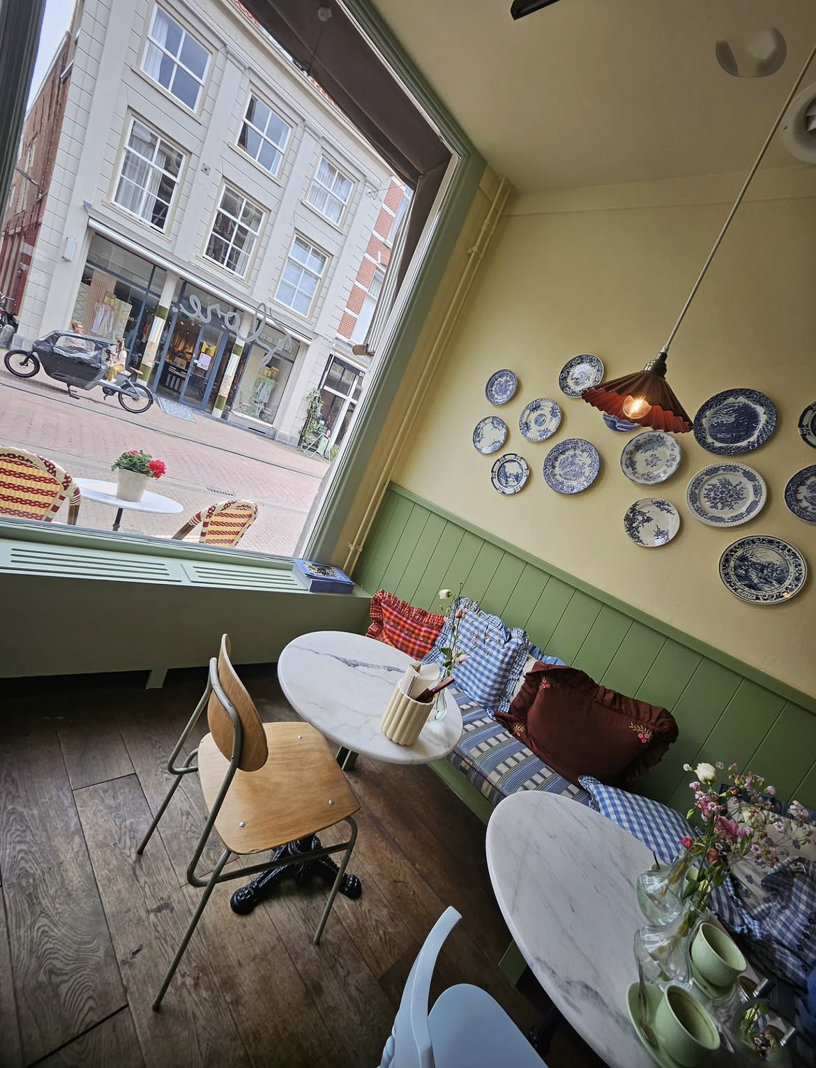 Binnenkant van een gezellige café met witte marmeren tafels, houten en plastic stoelen, bloemen en blauwe en witte servetten op banken met rode en blauw geruite kussens. Aan de muur hangende blauwe en witte keramische borden, grote raam met uitzicht op straat met winkels en fietsen.