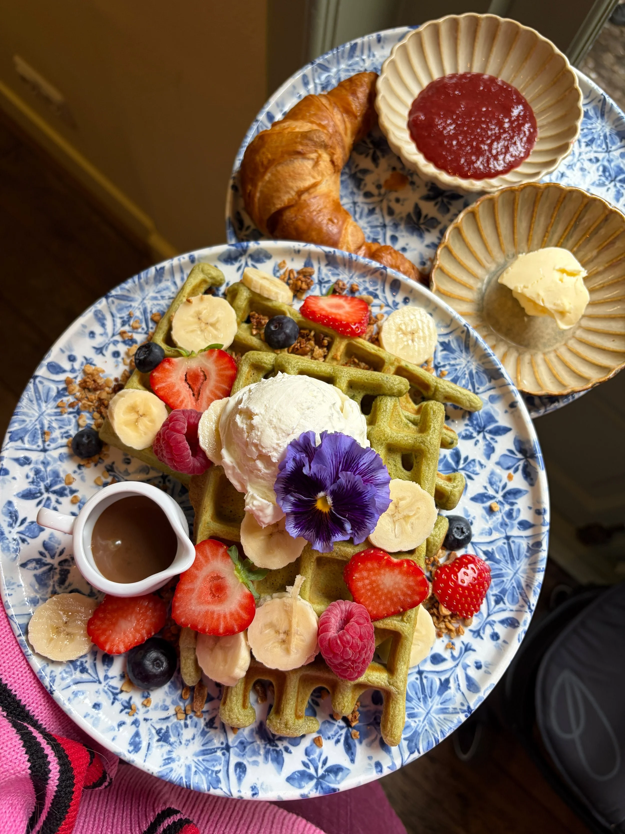 Een bord met wafels bedekt met vers fruit zoals aardbeien, blauwe bessen, bananen, en gevuld met ijs. Ook staan er kleine schaaltjes met jam, boter, en een kopje koffie.