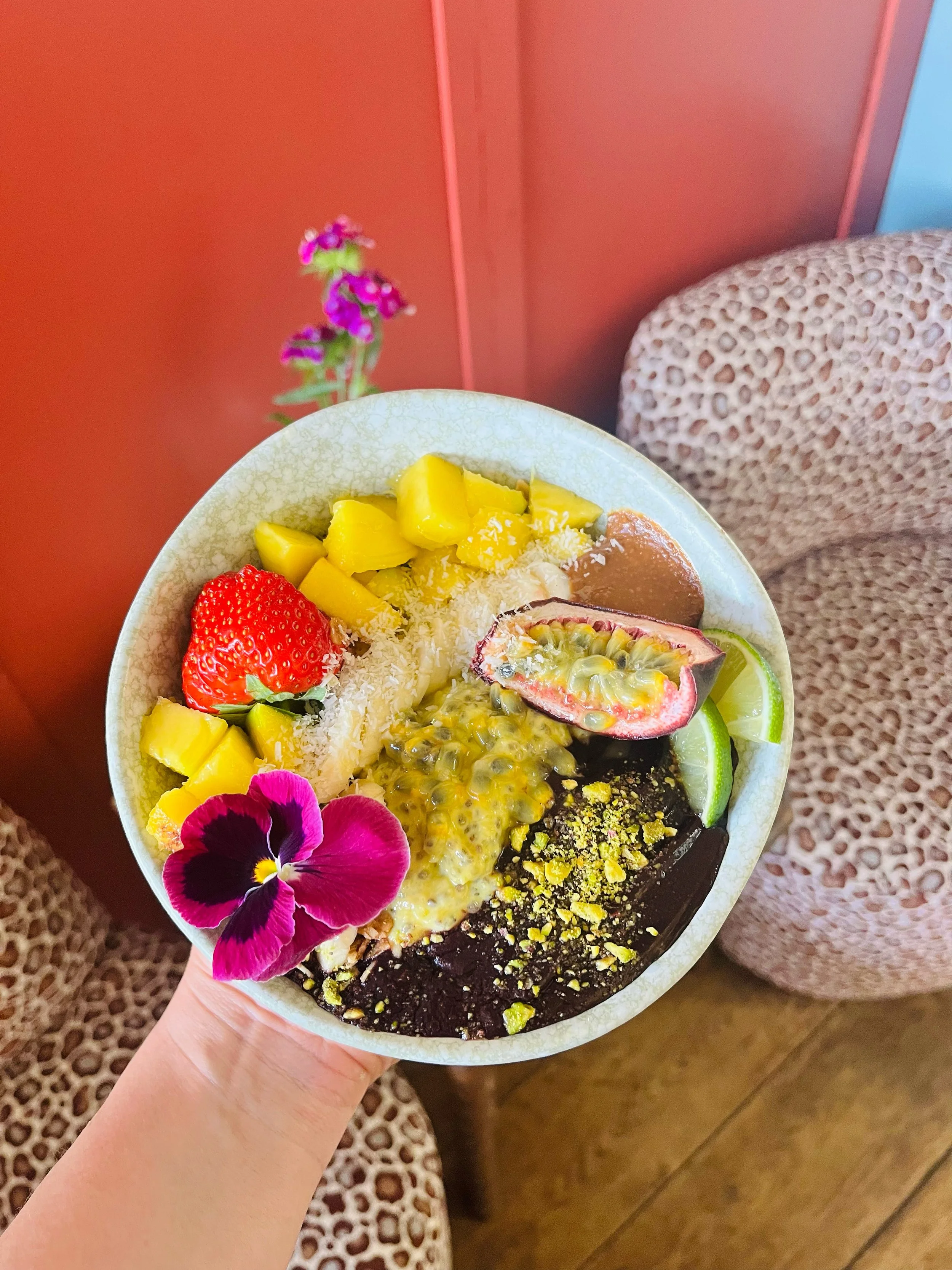 Bowl met diverse kleurrijke fruitsoorten en bloemen, inclusief aardbei, passievrucht, mango, limoen en viooltje, gehouden door een persoon.