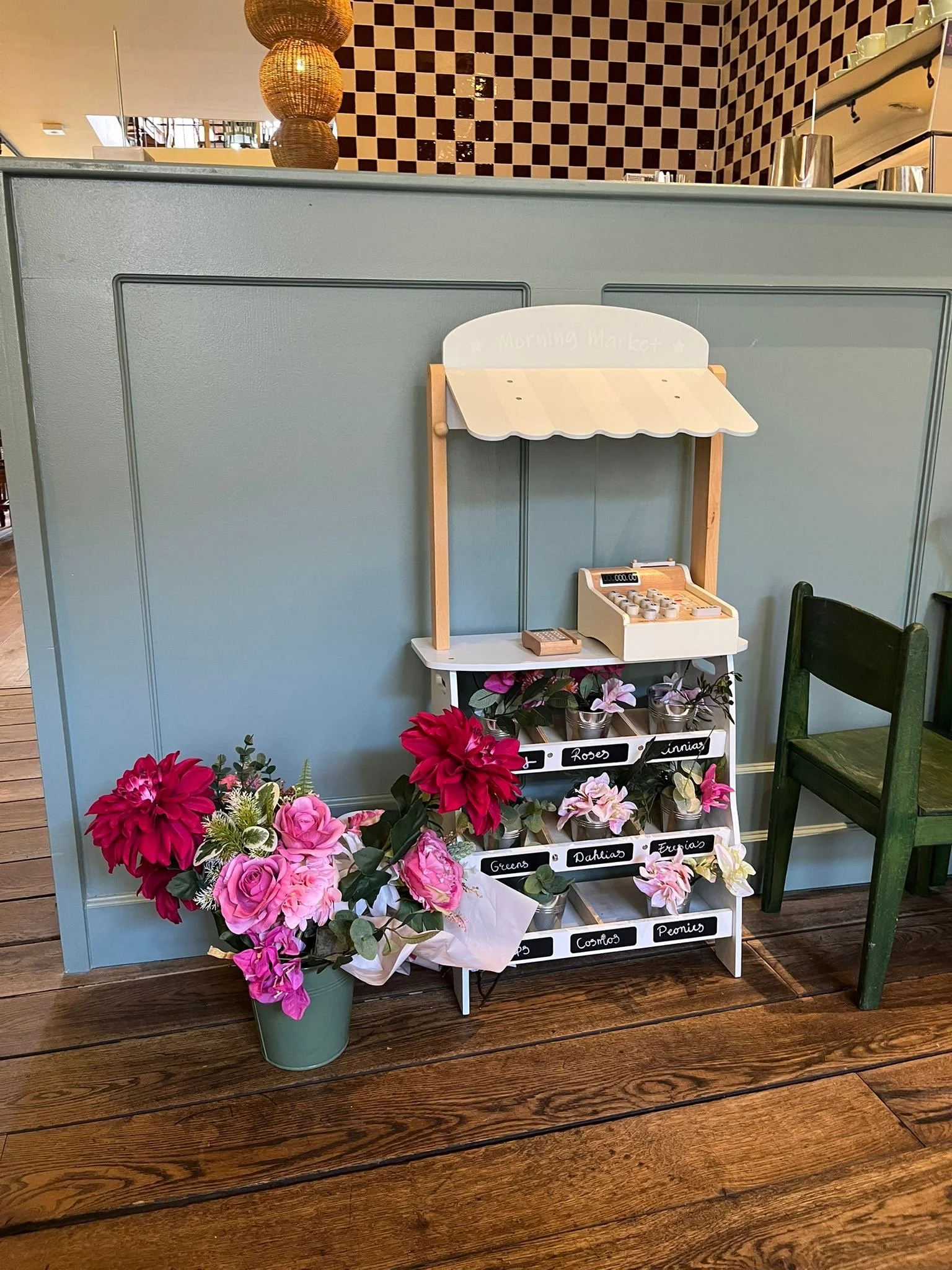 Een groene bloempot met roze en paarse bloemen, een witte houten speelgoedkassa, een witte winkelrek met labels voor verschillende bloemsoorten en een groene stoel. De achtergrond bestaat uit een blauwe muur en een donkerhouten vloer.