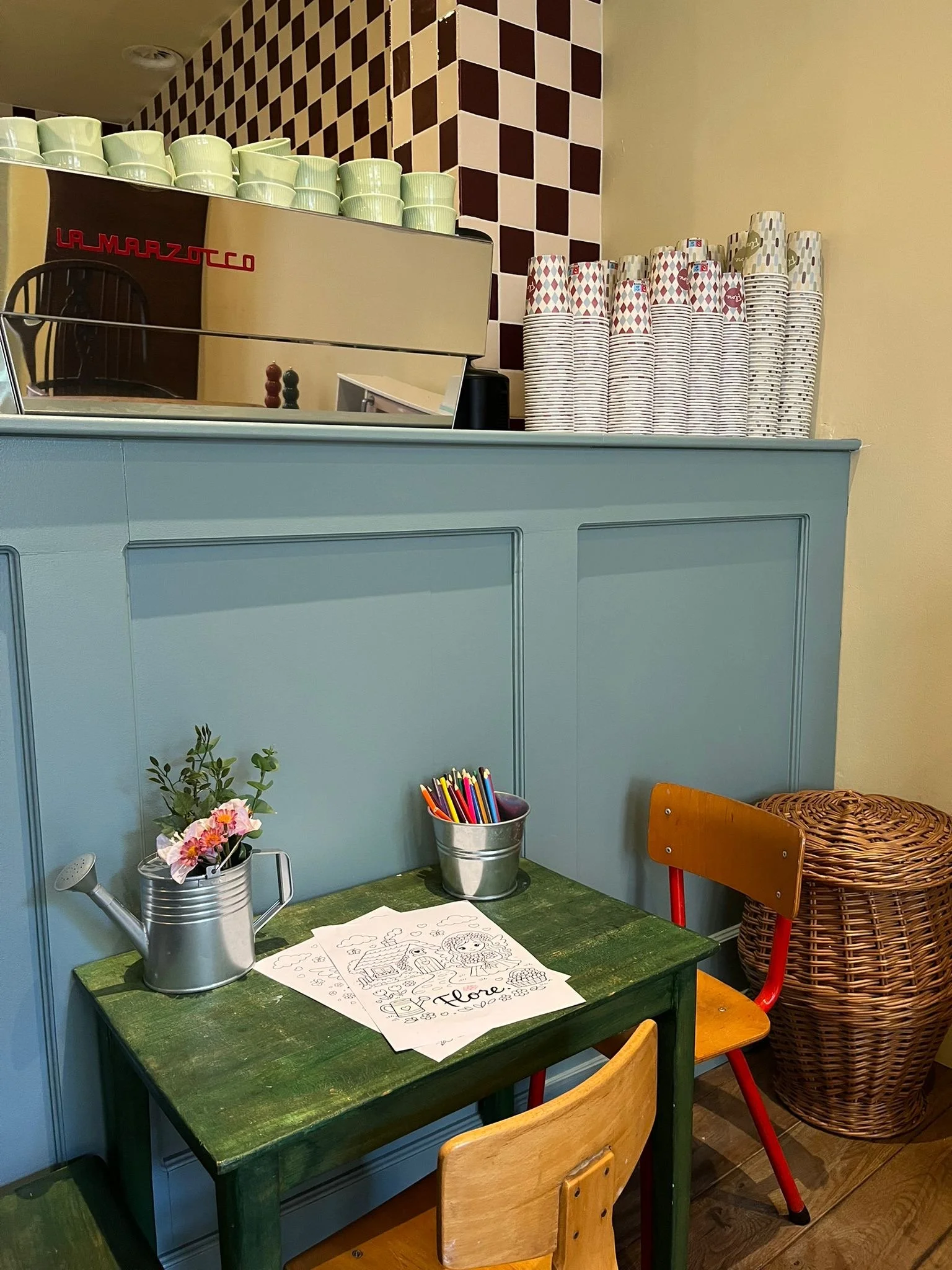 Een groene houten tafel met kleurpotloden in een metalen kan, een kleurboek met tekeningen, een waterkan met bloemen, een kussen en een rieten mand naast een blauwe muur en stoelen.