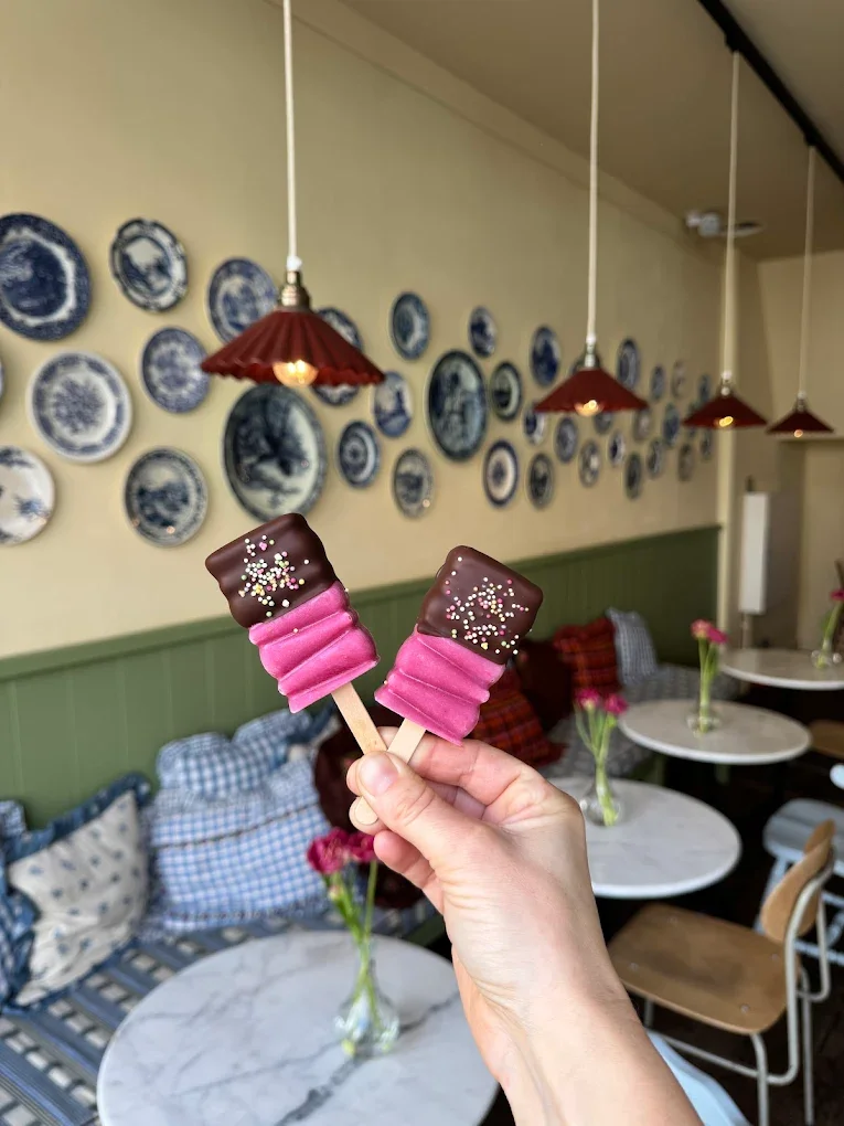 Een hand houdt twee ijsjes met een roze en chocolade topping in een gezellige, sfeervolle ruimte met blauwe en witte borden aan de muur, rondtafels met bloemen, en hanglampen.