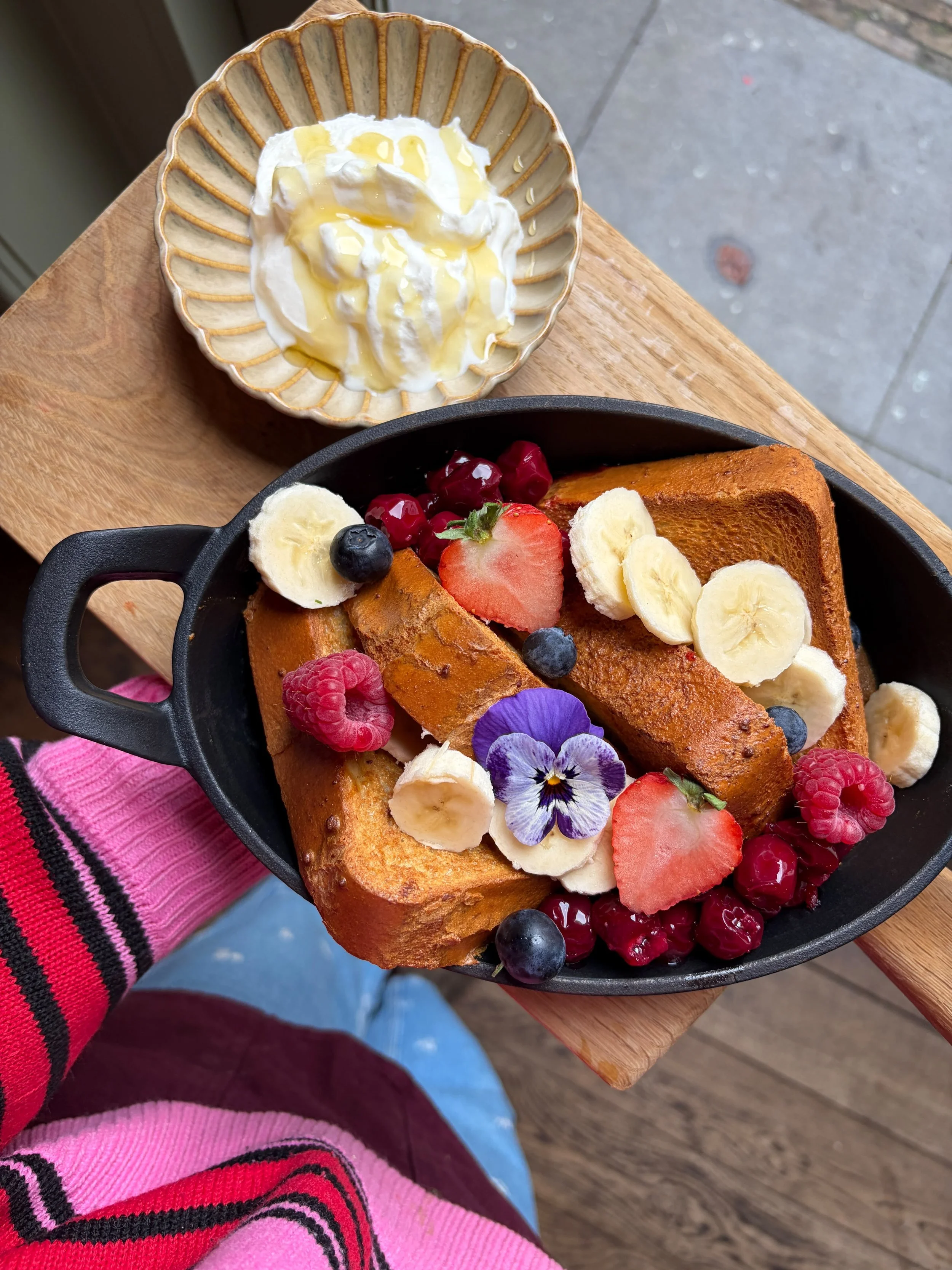 Paasbrood met banaan, aardbeien, blauwe bessen, frambozen en viooltjes, naast een schaaltje met yoghurt en honing, op een houten tafel.