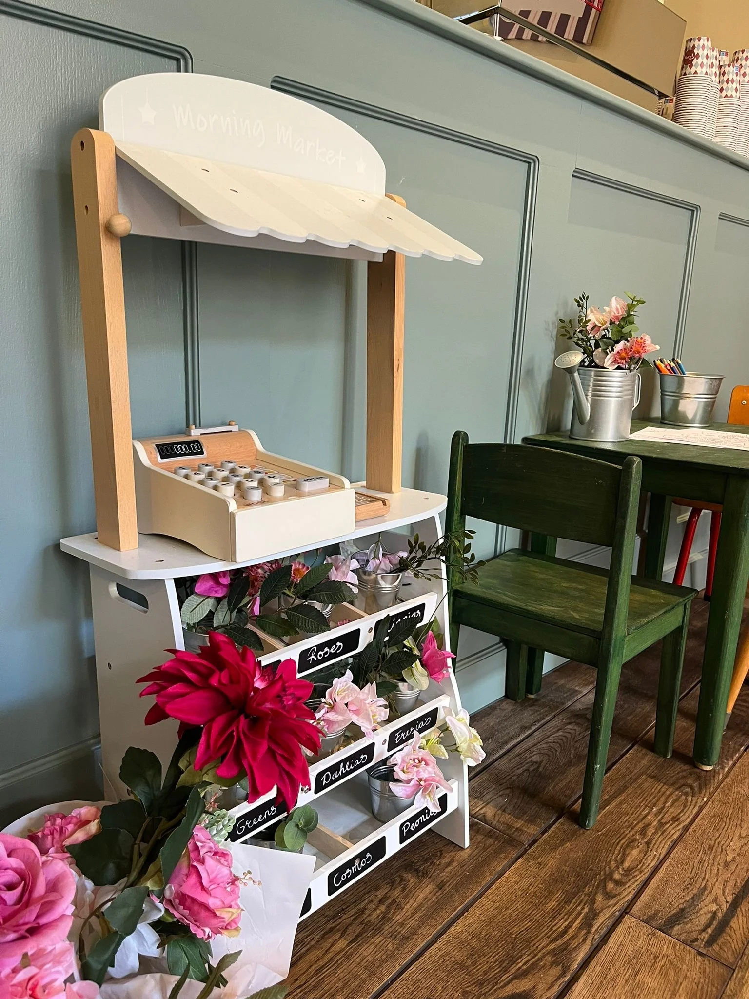 Een witte houten speelgoedkassa met een digitaal display en klinken, naast een witte driehoekige stand met bloemen. Achter de speelgoedkassa een grijze muur en een houten vloer.