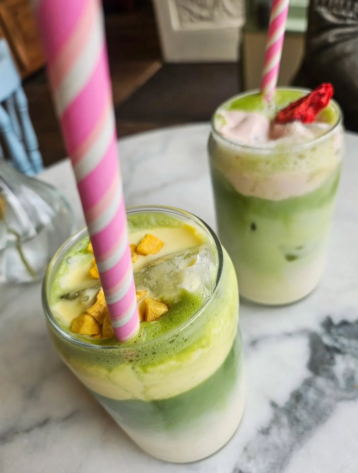 Twee groene en witte milkshakes met roze en witte rietjes en fruit en cruesli als topping.