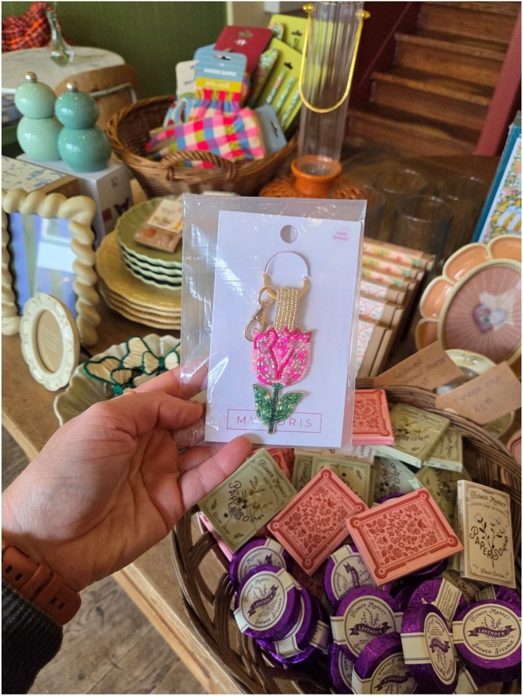 Hand holding a bag with a pink and green embroidered tulip keychain in a store with various decorative items and soaps on display.