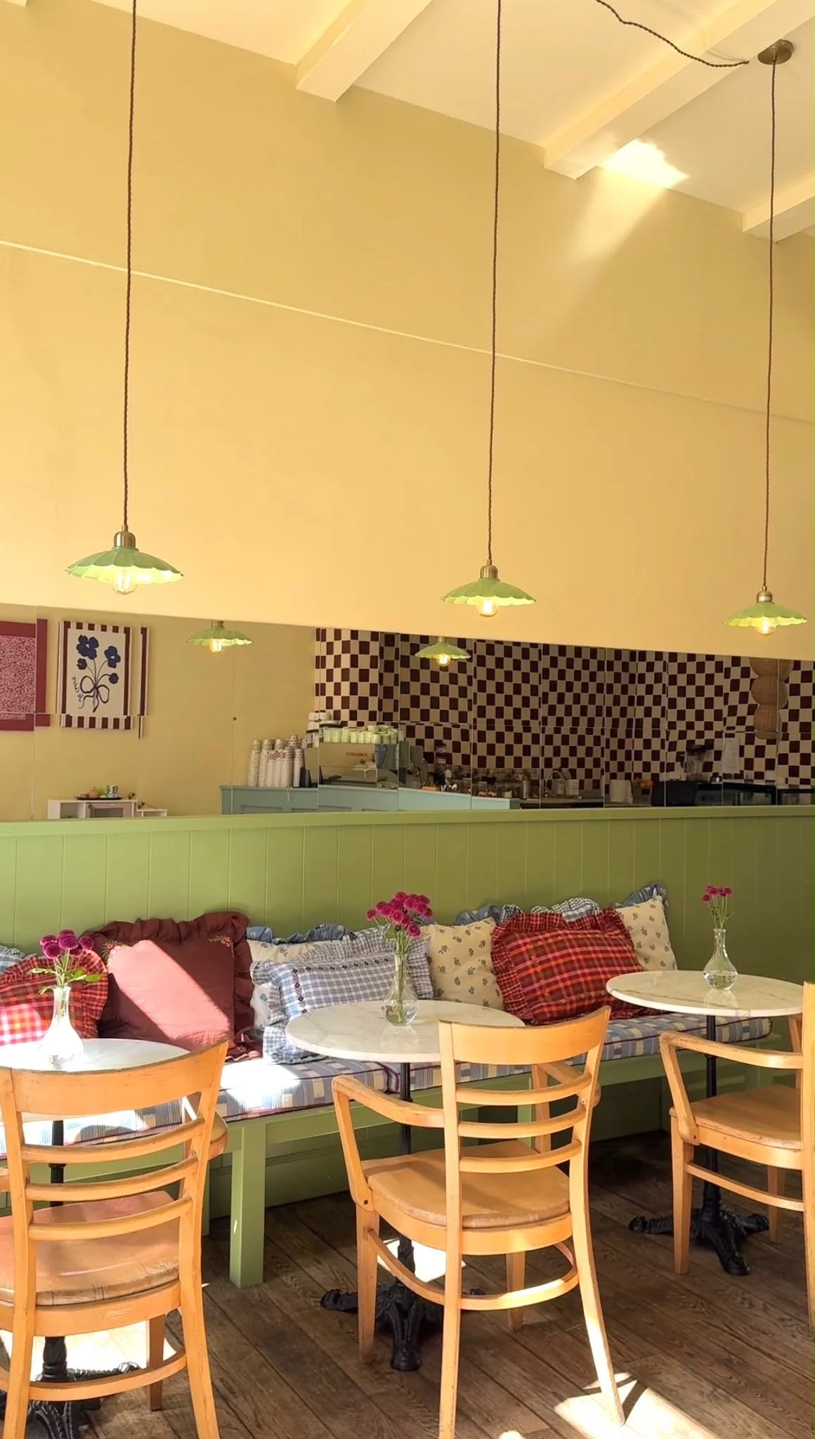 Interieur van een gezellige restaurant of café met geelgroene wand, houten stoelen, en tafels met vazen met bloemen.