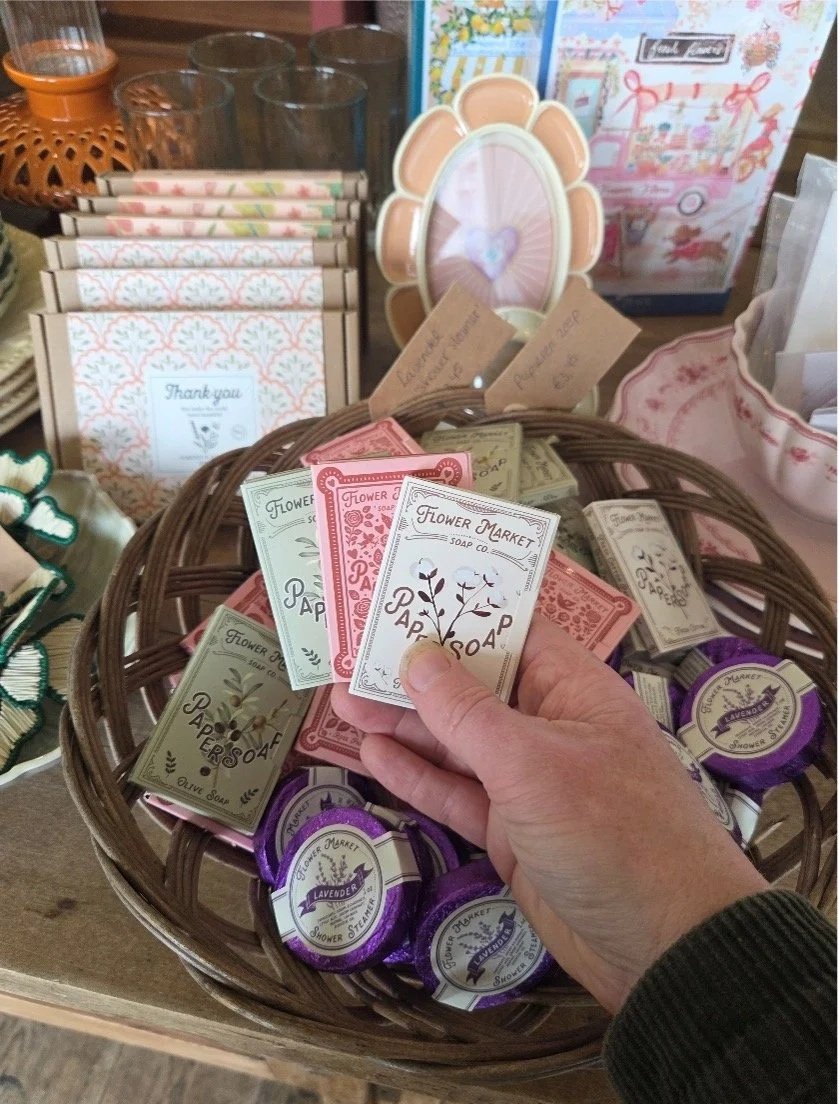 Een hand die een pakje zeep van bloemenmarkt vasthoudt, met andere verpakkingen zeep en winkelartikelen op de achtergrond.