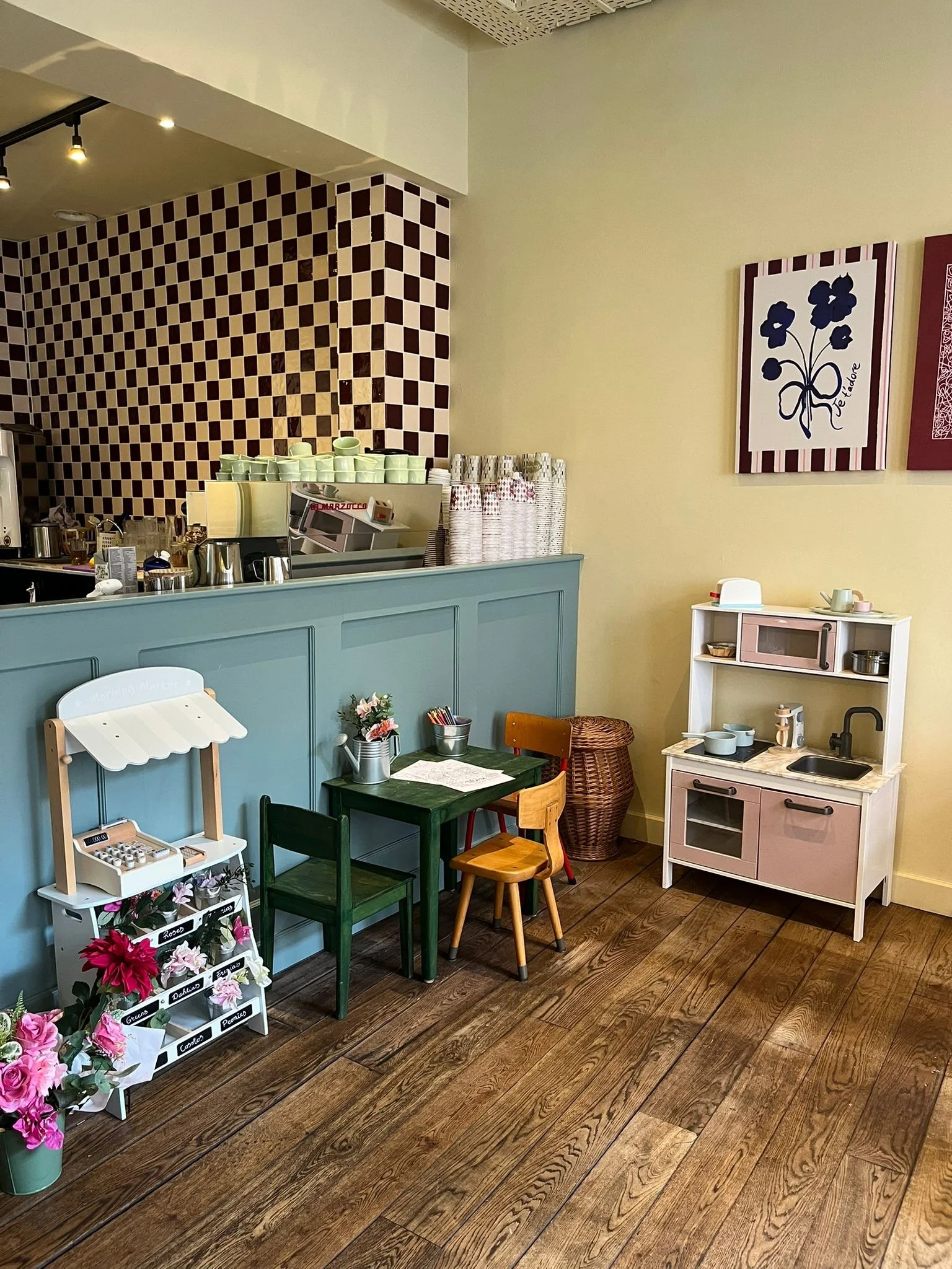 Kleine speelhoek in een speelhoek met houten vloer, groene en houten kinderstoelen, een witte speelgoedkeuken, bloemen, een speeltoreraam, en decoratie op de muur. De muur heeft een schildering van blauwe bloemen met de tekst "Je favoriete".