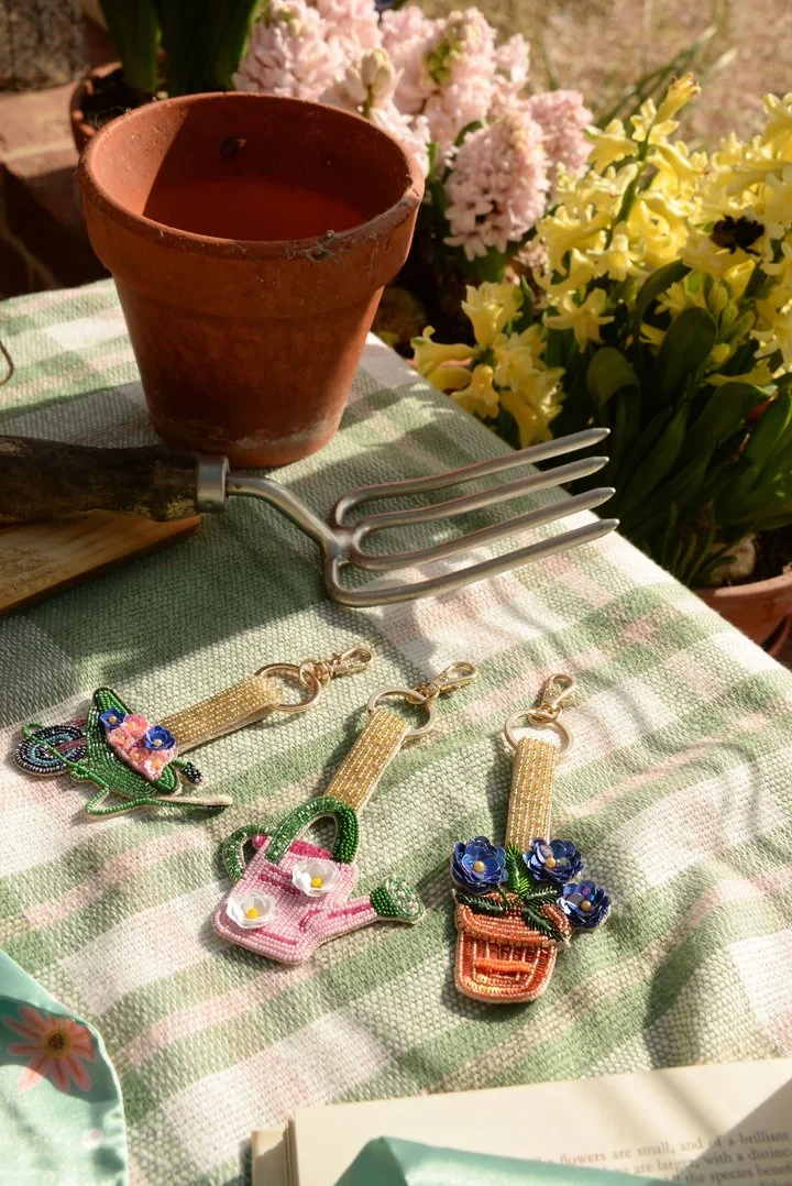 Twee emblemen van geëmbroide textiel, een met een vis en een ander met een bloem, liggen op een groene en witte geruite tafelkleed, met potten met gekleurde bloembollen en een tuinfork op de achtergrond.