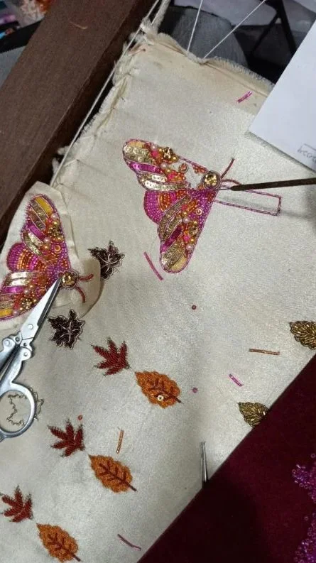 Een lap Stof met geborduurde vlinders en bladeren, waarbij een kunstenaar met pincet en pincet aan het borduren is