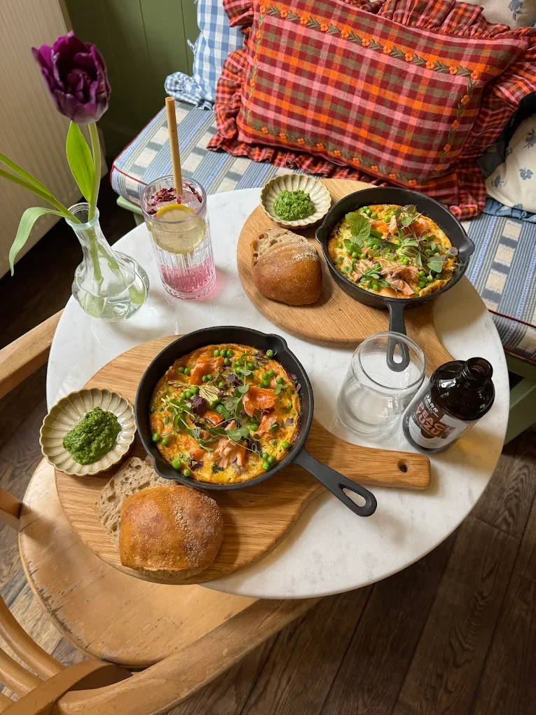 Twee hartige pannenkoeken met vis, groene peas en microgroenten, geserveerd in kleine gietijzeren pannen op houten schotels, vergezeld van brood, een potje groene saus, en een drinkglas met drankje op een wit marmeren tafel met een bloem in een vaas.