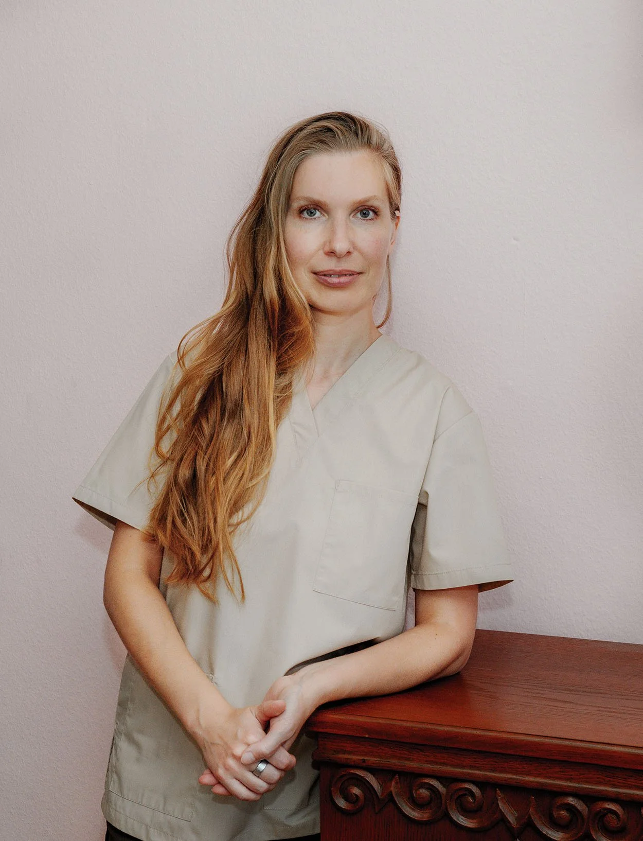 Eine Frau mit roten Haaren in einer weißen Arbeitsschürze lehnt an einer Holzkommode vor einer weißen Wand.