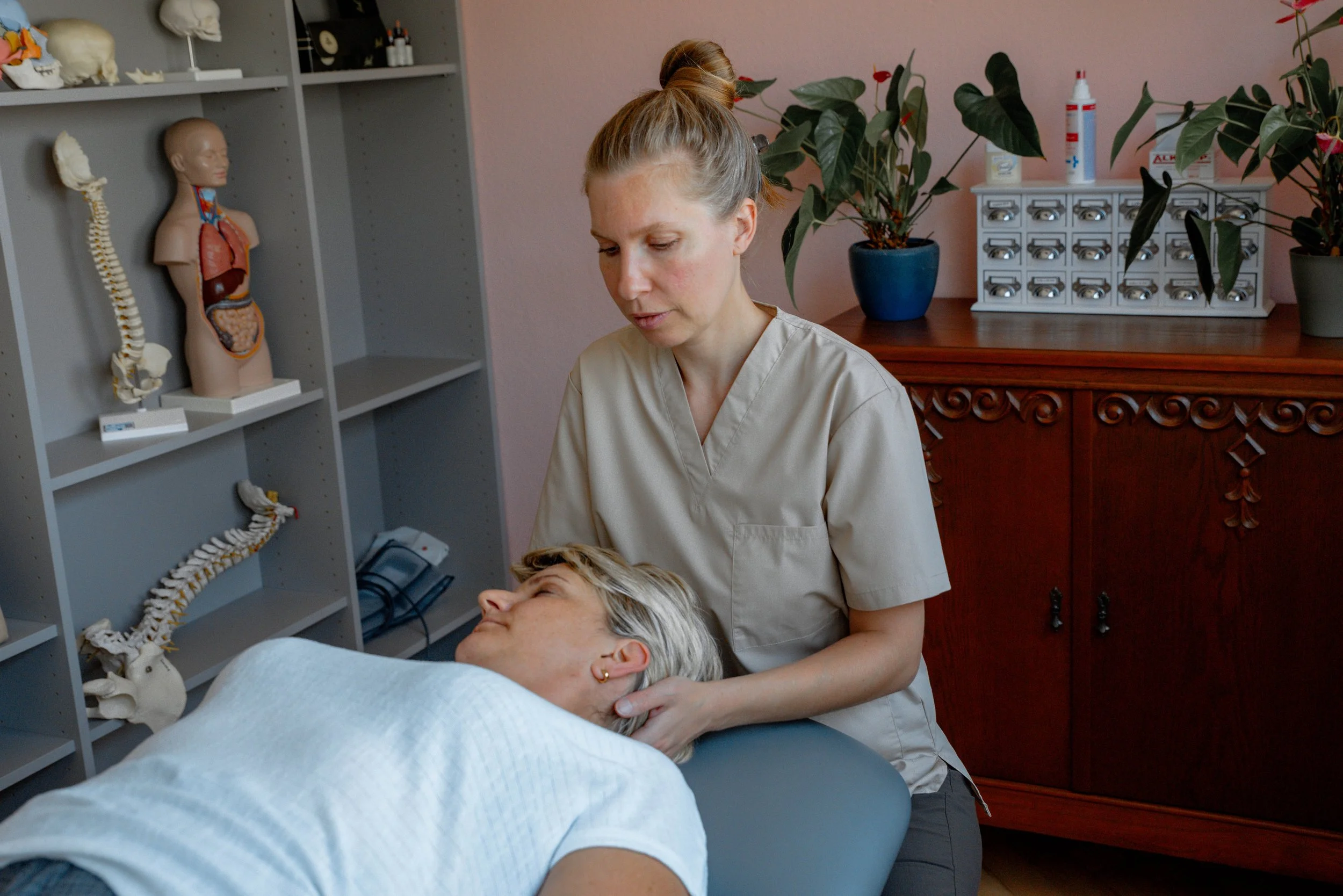 Eine medizinische Fachkraft kümmert sich um eine ältere Patientin im Behandlungszimmer, mit anatomischen Modellen im Hintergrund.