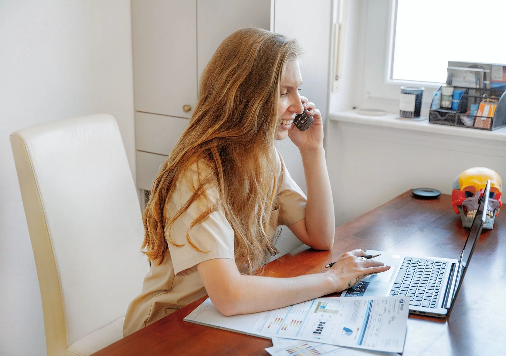 Frau mit langen roten Haaren arbeitet am Computer, telefoniert, sitzt an einem Schreibtisch mit Dokumenten, einem geköpften menschlichen Schädel mit bunten Farben und Skizzen, im Hintergrund eine Fenster und ein Regal.