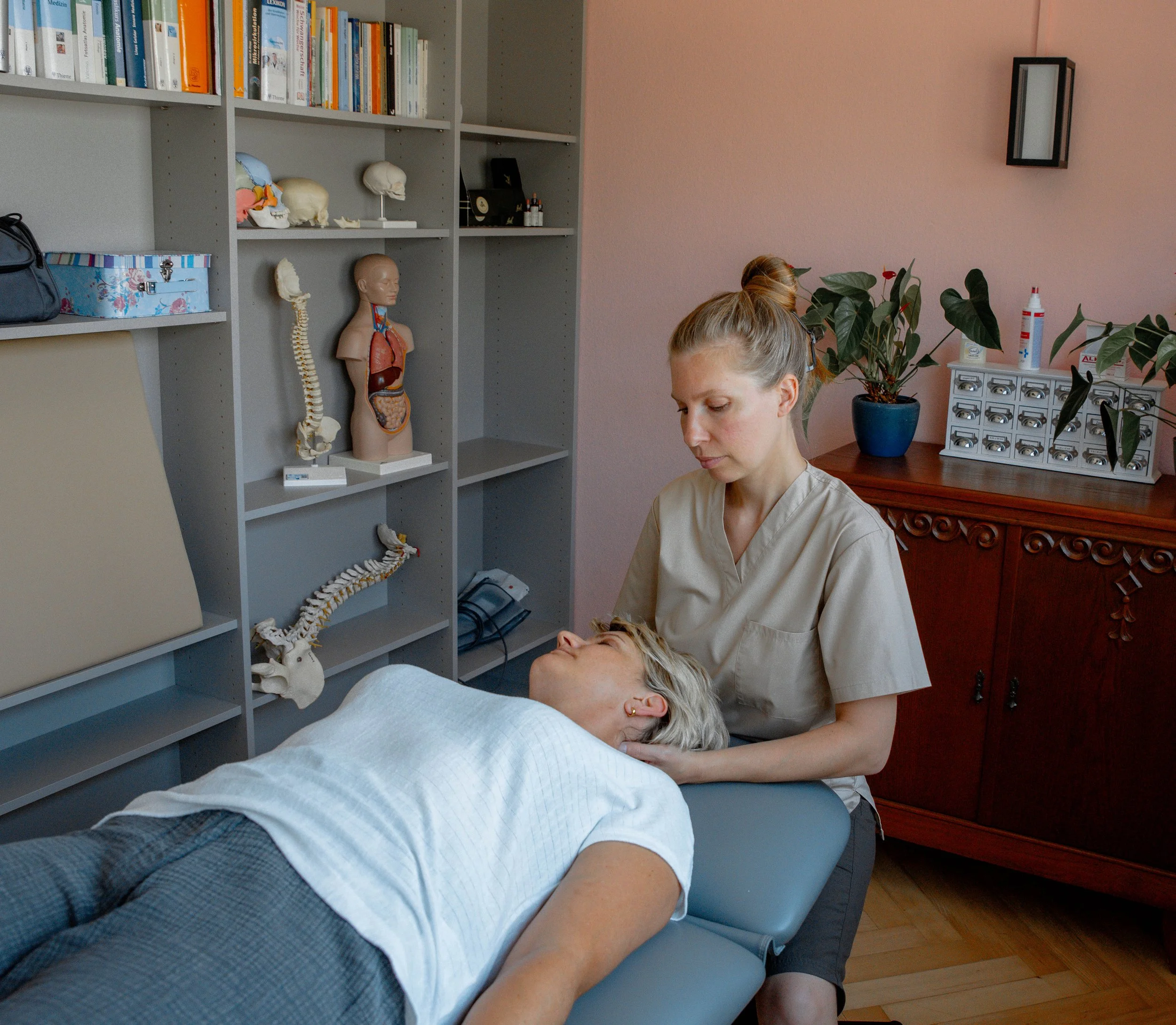 Eine Person liegt auf einer Couch und ein Therapeut oder eine Therapeutin führt eine Behandlung durch, möglicherweise eine physiotherapeutische Massage oder Chiropraktik. Im Hintergrund sind Bücherregale mit medizinischen und anatomischen Büchern sowie verschiedene medizinische Modelle und Gegenstände zu sehen.