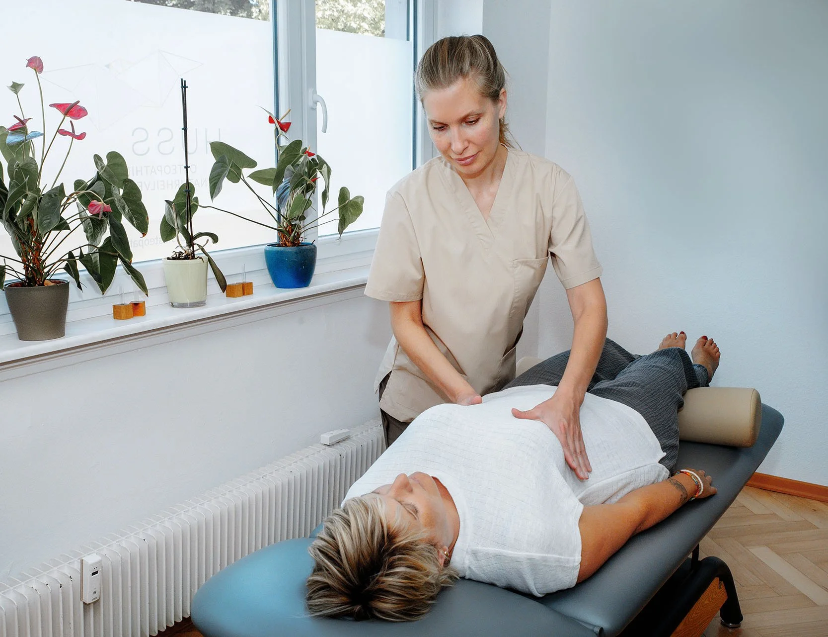 Eine Physiotherapeutin behandelt eine Patientin auf einer Behandlungsliege in einer Praxis. Im Hintergrund sind Fenster mit Pflanzen auf der Fensterbank.