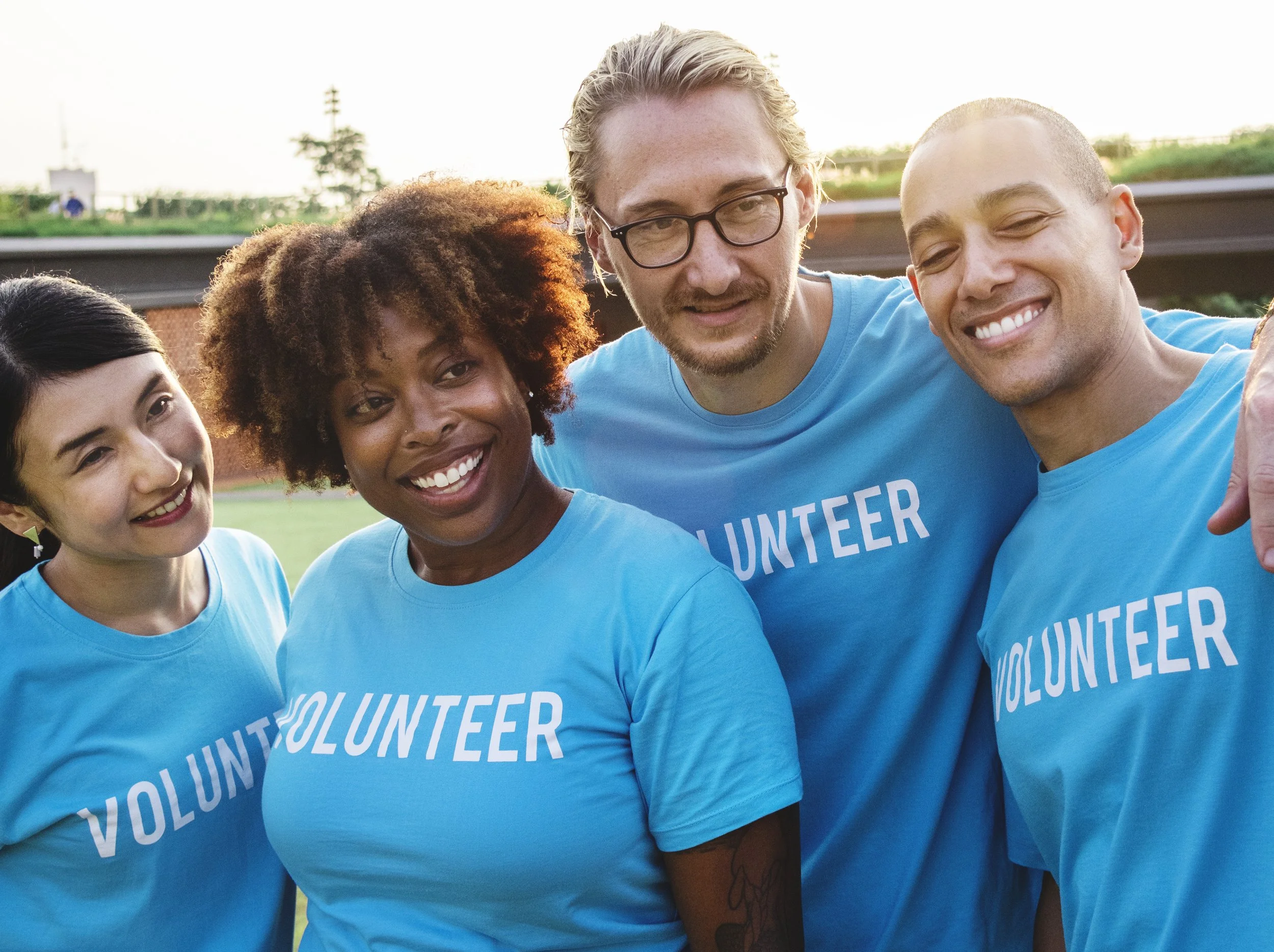 Group photo of diverse volunteers in blue shirts with 'VOLUNTEER' printed on them, smiling at an outdoor event with a park and trees in the background.