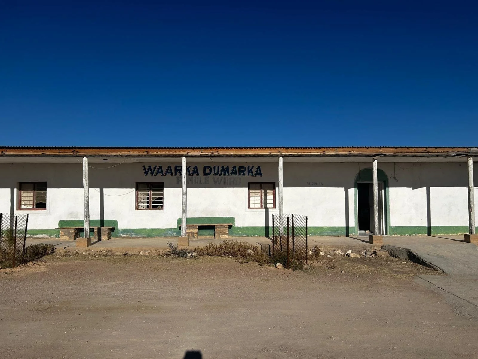 A single-story building with a sign that reads 'WAARKA DUMARKA' under a clear blue sky. The building has three windows with bars, two benches outside, and a main entrance door. The exterior walls are painted white with a green trim near the ground, and the roof is corrugated metal supported by wooden poles.