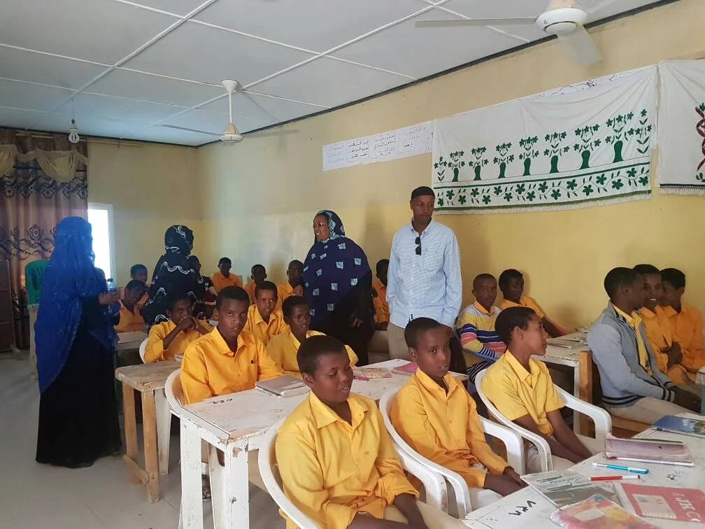 Classroom with students in yellow uniforms, two women standing, a man standing, and educational posters on the wall.