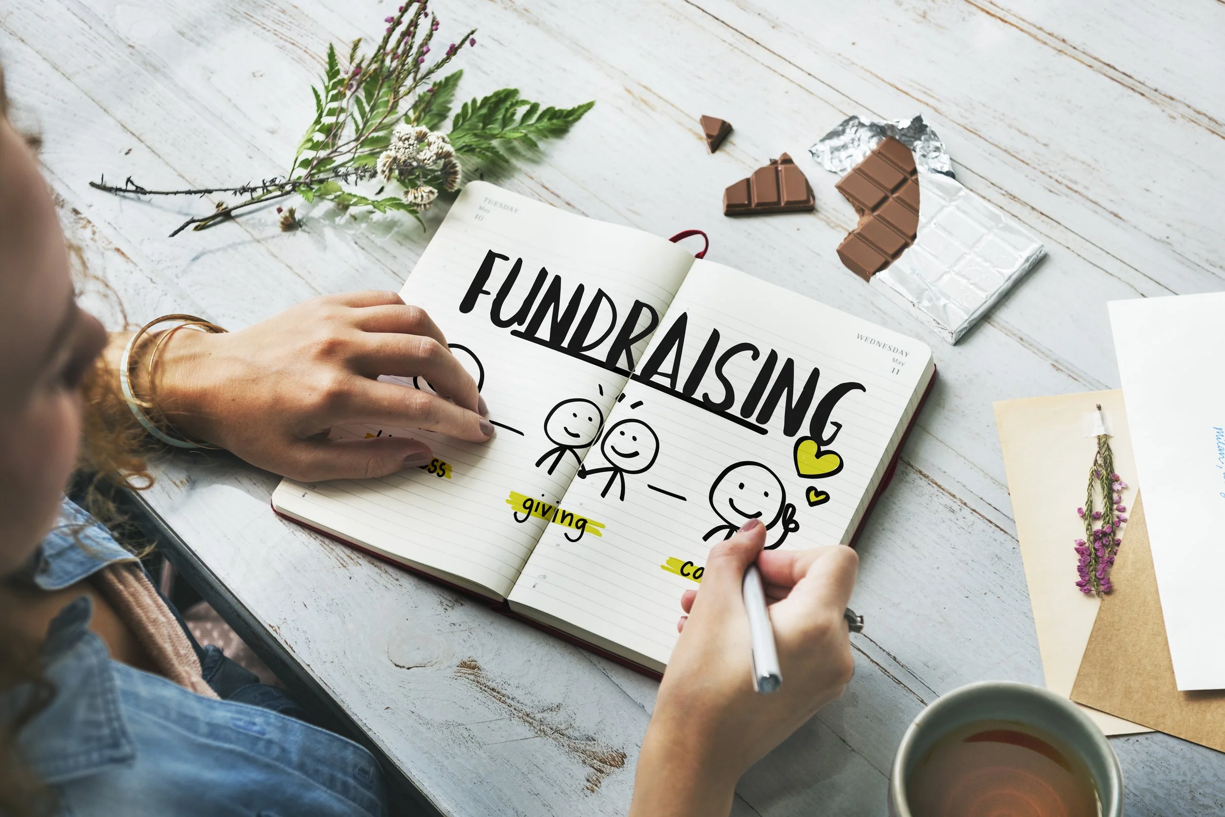 A person is drawing a cartoon figure of two smiling stick figures holding hands on a lined notebook page that has the word 'FUNDRAISING' written at the top. There are sections labeled 'giving' and 'calling' highlighted in yellow. The person is sitting at a white wooden table with a partially unwrapped chocolate bar, a small flower arrangement, and several photos or notes nearby. A mug of tea or coffee sits at the bottom of the image.